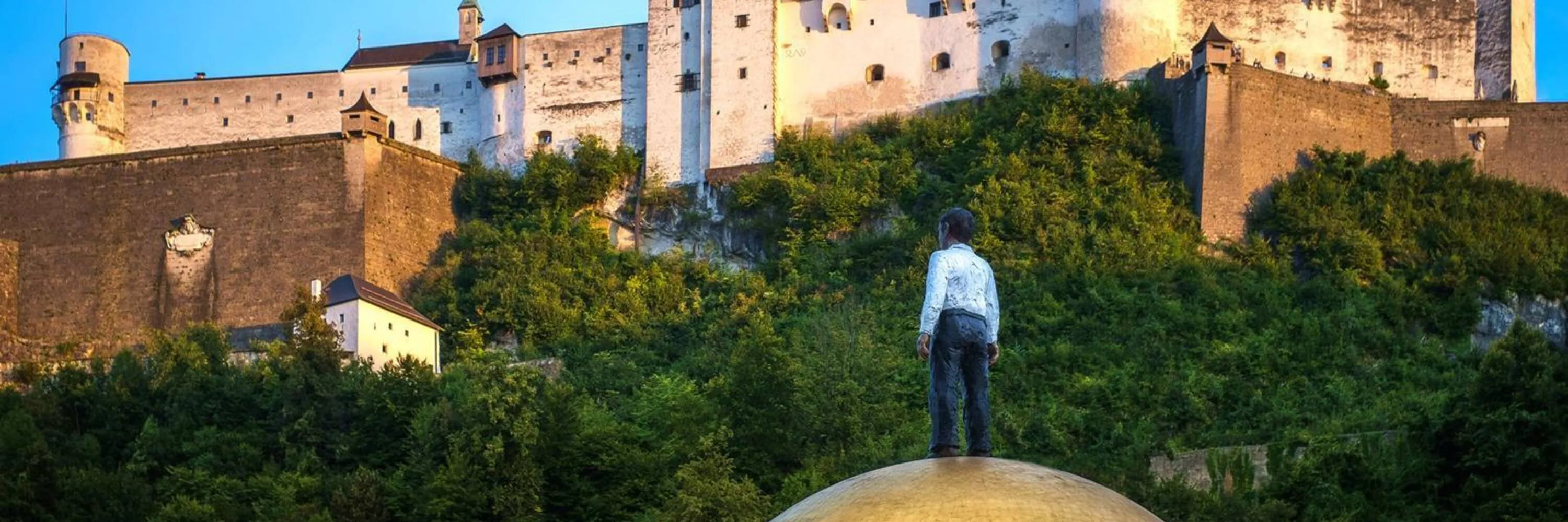 Un hombre está de pie sobre una gran escultura de una esfera dorada, mirando hacia la fortaleza histórica iluminada en una colina verde al atardecer.