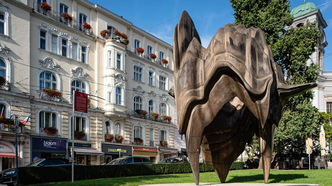 Gran escultura abstracta de bronce que se asemeja a una forma orgánica parecida a una concha, situada en una mediana cubierta de hierba frente a edificios históricos de Salzburgo con balcones adornados con flores bajo un cielo azul