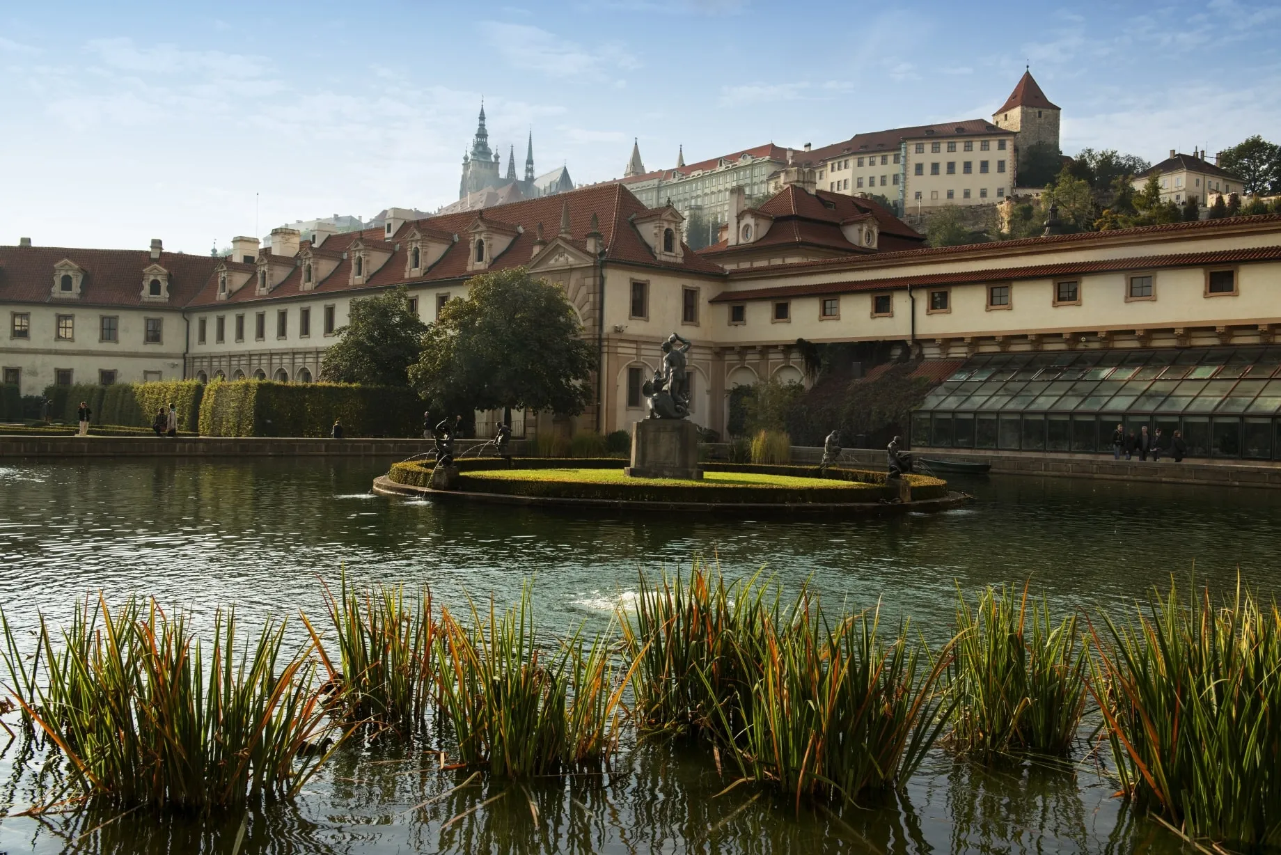 Staw z centralną fontanną i posągiem w Ogrodach Pałacu Waldstein, z historycznymi budynkami i zamkiem w tle.