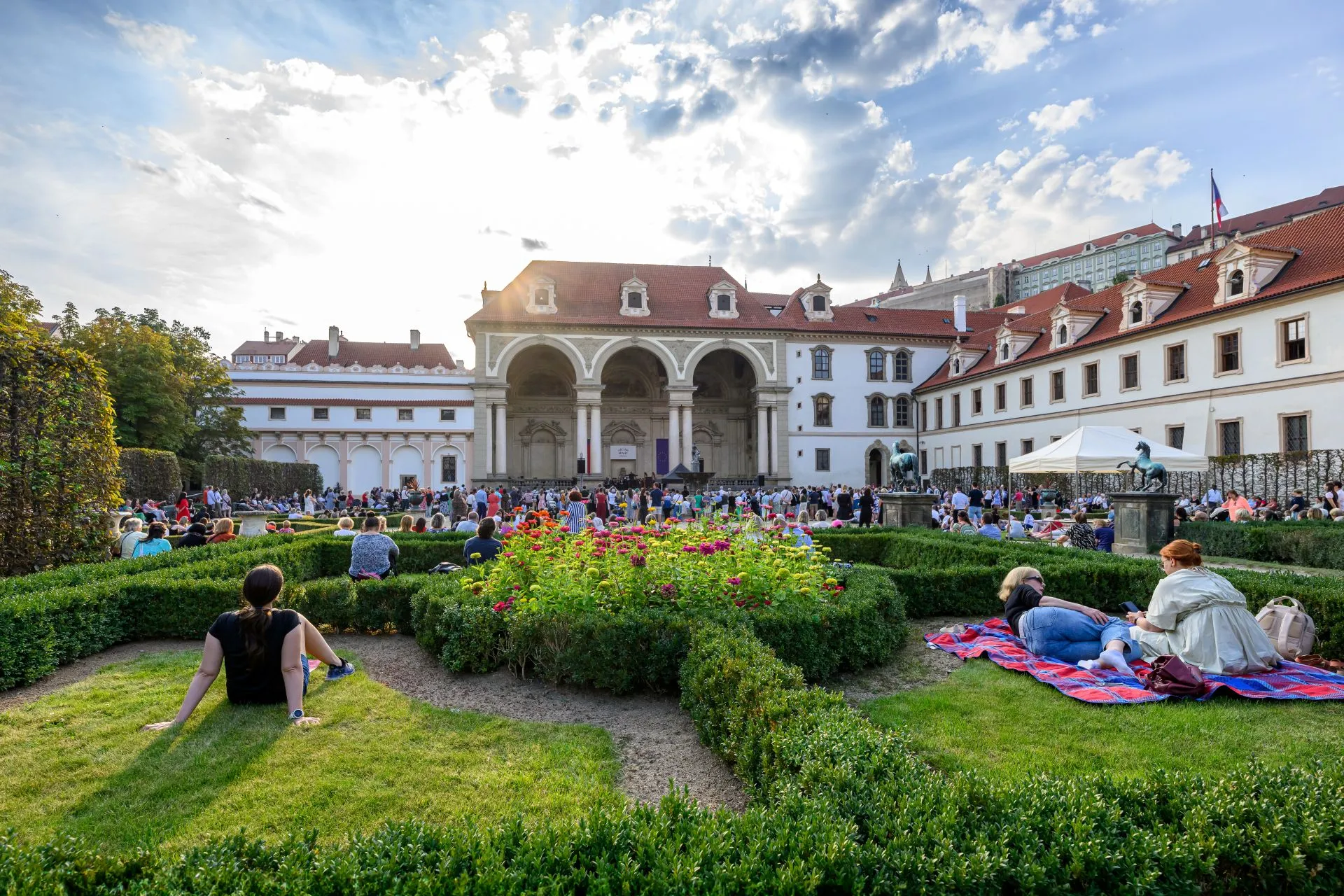 Ludzie relaksujący się na trawie w barokowym ogrodzie z wypielęgnowanymi żywopłotami, rabatami kwiatowymi i dużym białym budynkiem z arkadowymi kolumnadami w tle.