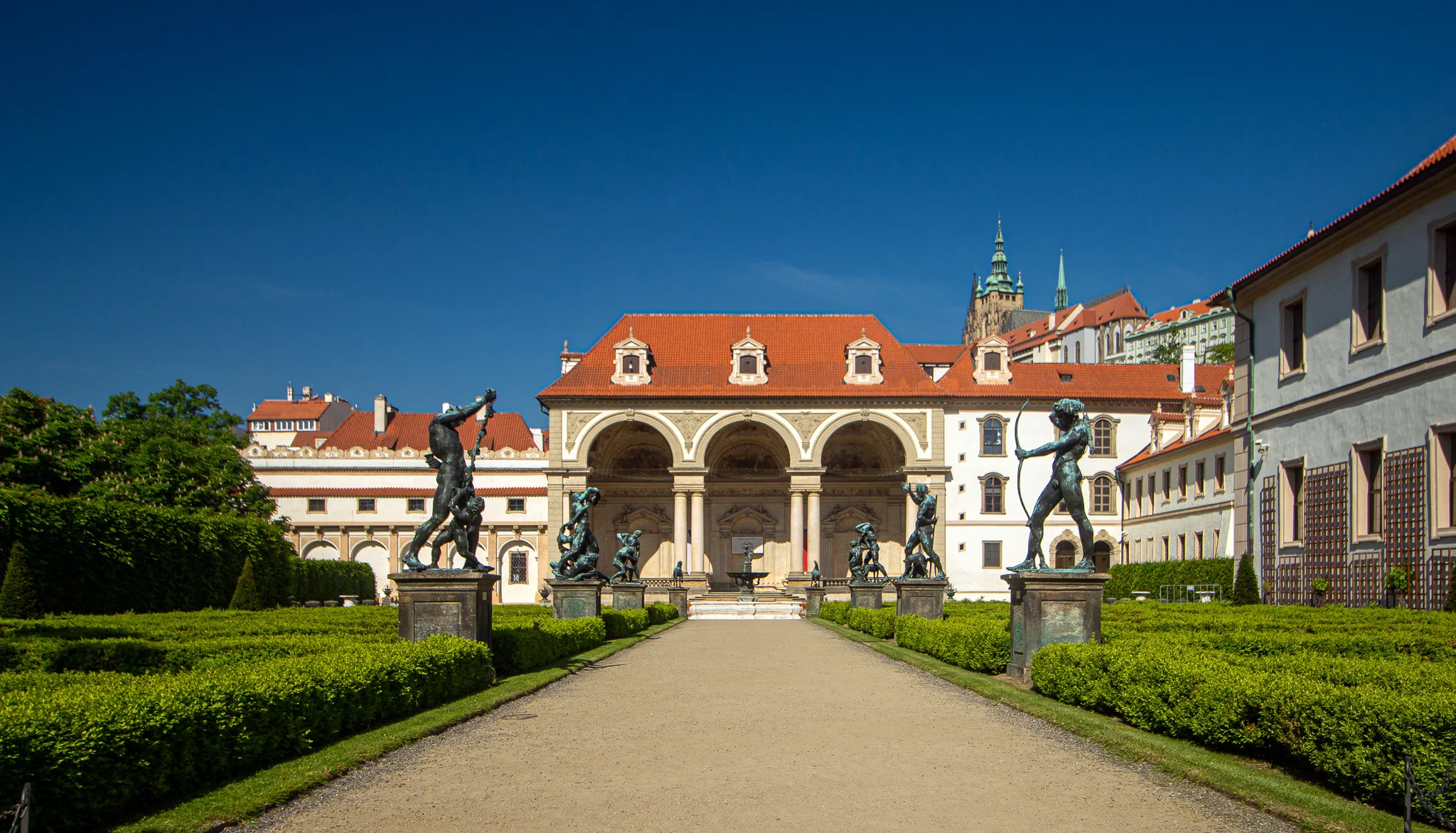 Szeroka ścieżka w formalnym ogrodzie prowadzi do okazałego budynku z dachem pokrytym czerwonymi dachówkami i arkadową kolumnadą, otoczonego zielonymi żywopłotami i posągami pod czystym błękitnym niebem.