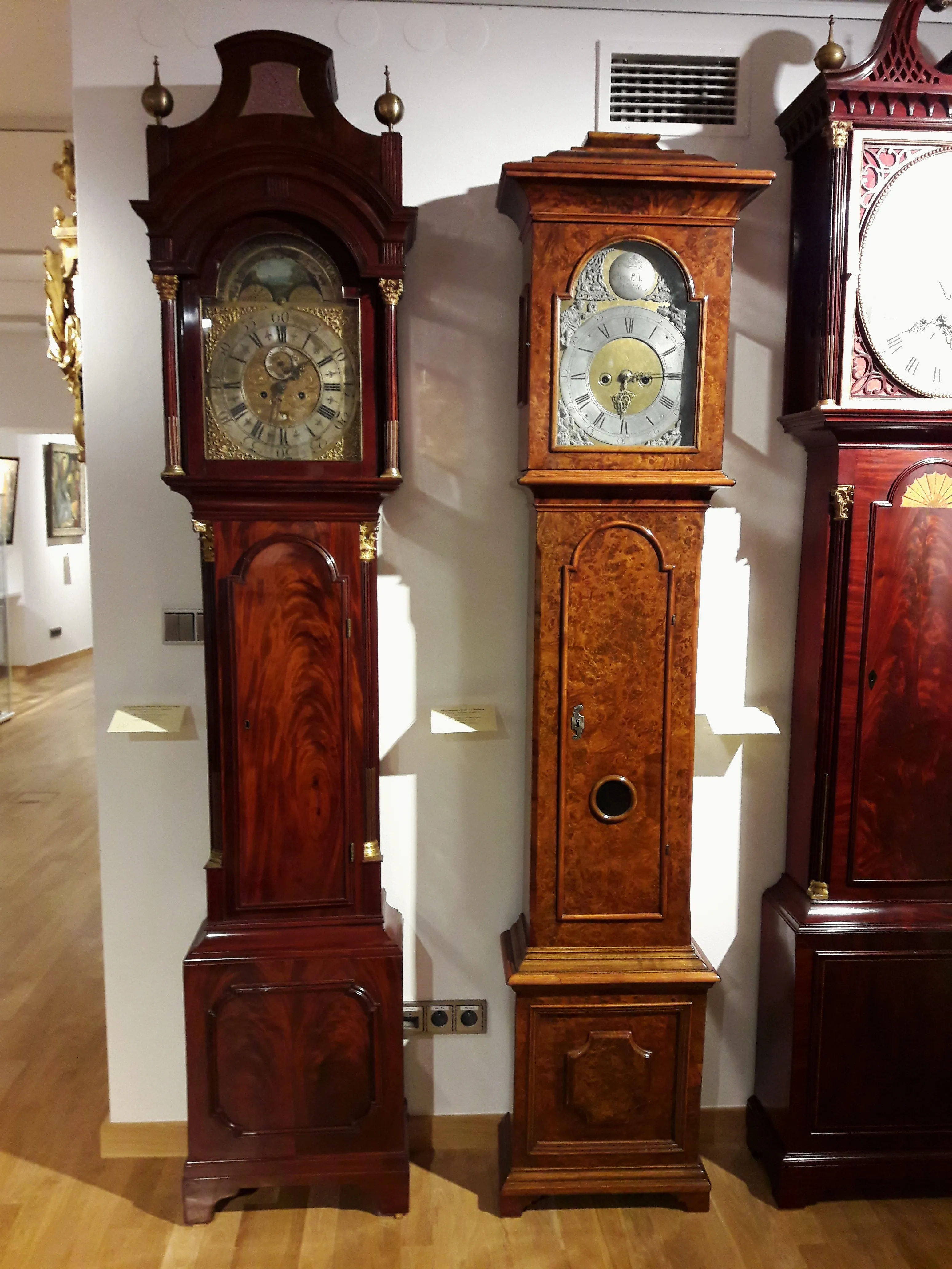 Drie staande klokken met sierlijke houten kasten en wijzerplaten tentoongesteld in een museumgalerij