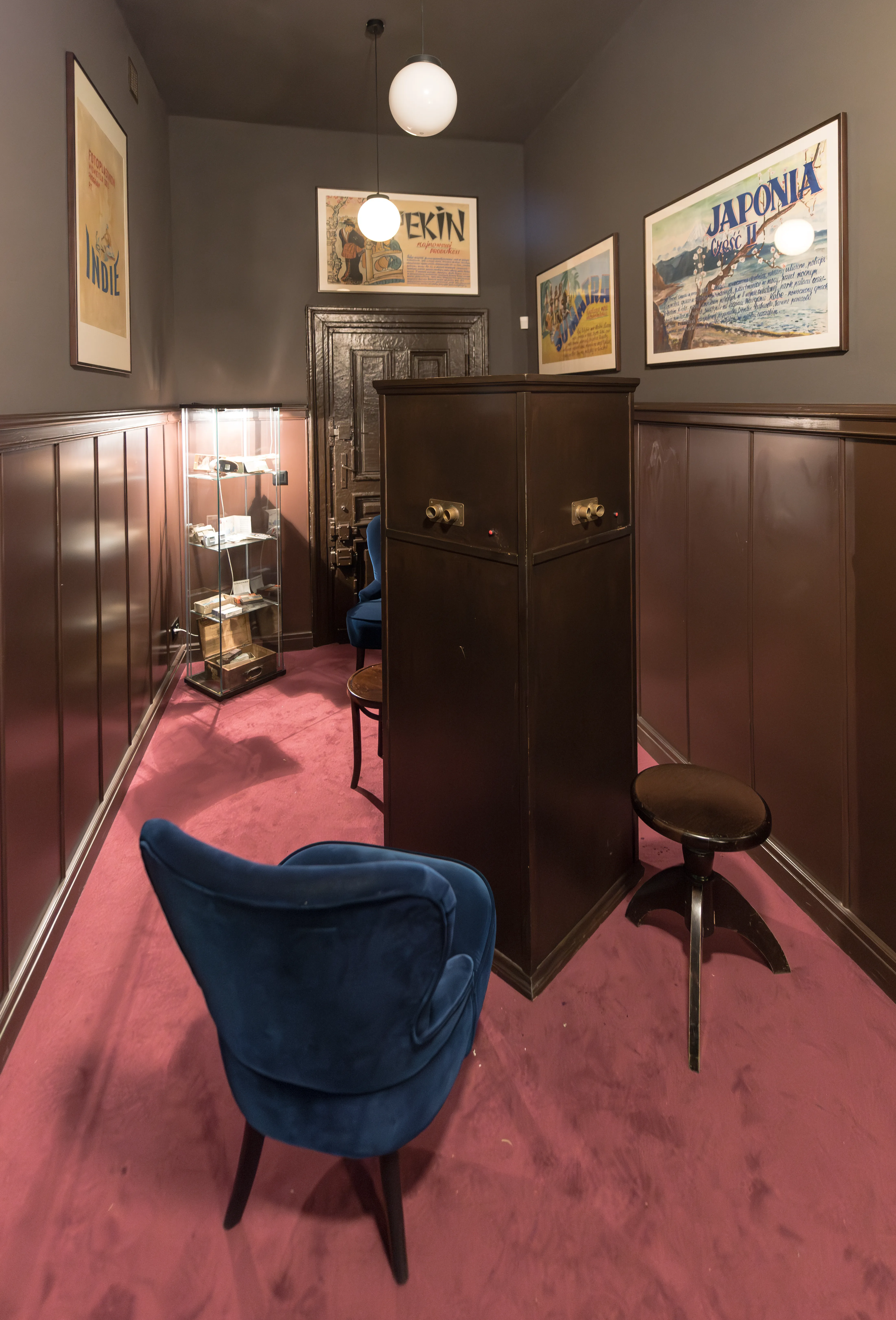 Interior del Fotoplastikon de Varsovia con paneles de madera oscura, alfombra rosa, silla de terciopelo azul y carteles vintage en las paredes