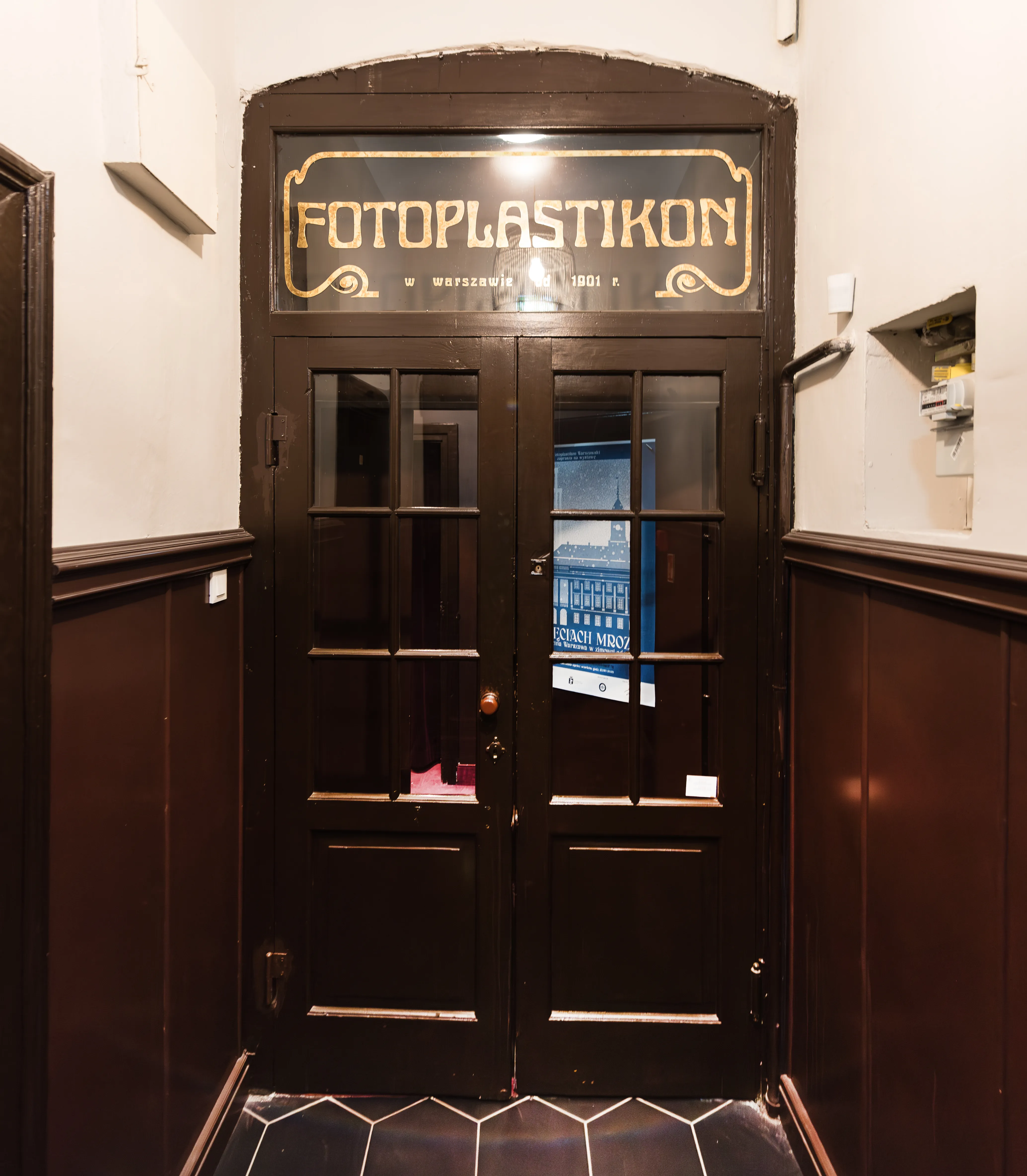Puerta de madera oscura con panel de vidrio y letras doradas que dicen 'FOTOPLASTIKON' encima, en un pasillo con paneles de madera oscura y baldosas hexagonales.