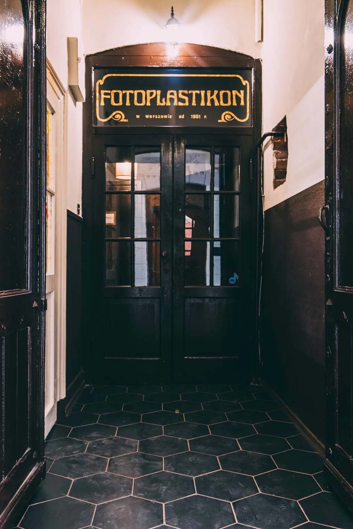 Puertas de madera oscura con paneles de vidrio debajo de un letrero vintage que dice 'FOTOPASTIKON' en letras doradas, en un pasillo con baldosas hexagonales negras.