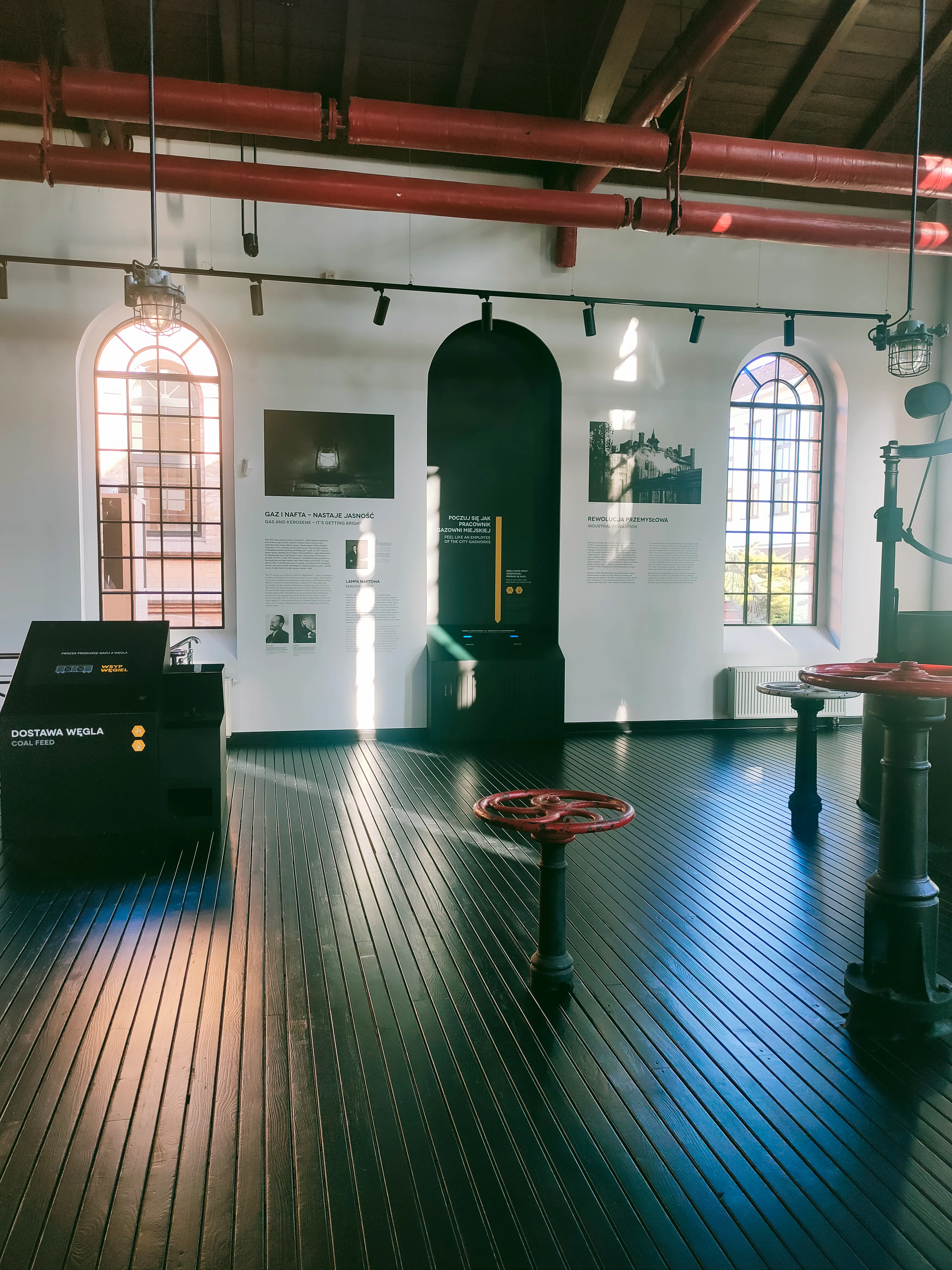 Interior of a museum with wooden floors, red pipes on the ceiling, arched windows, and exhibition displays