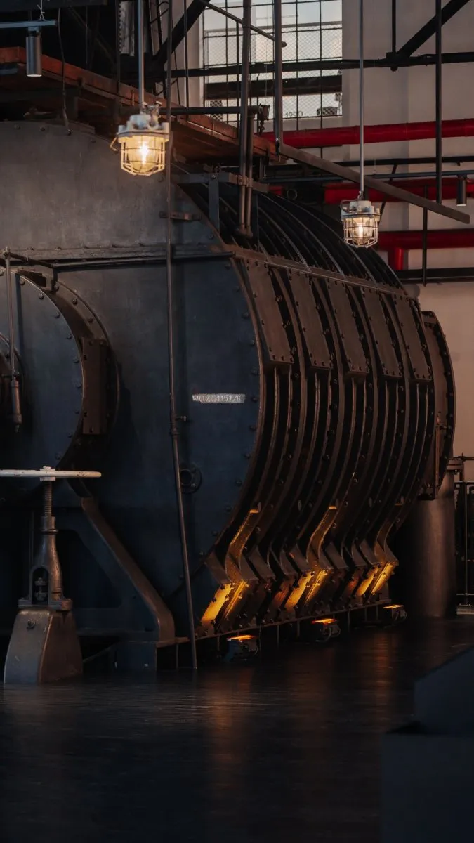 Large industrial boiler with ribbed metal casing and hanging pendant lights inside a museum