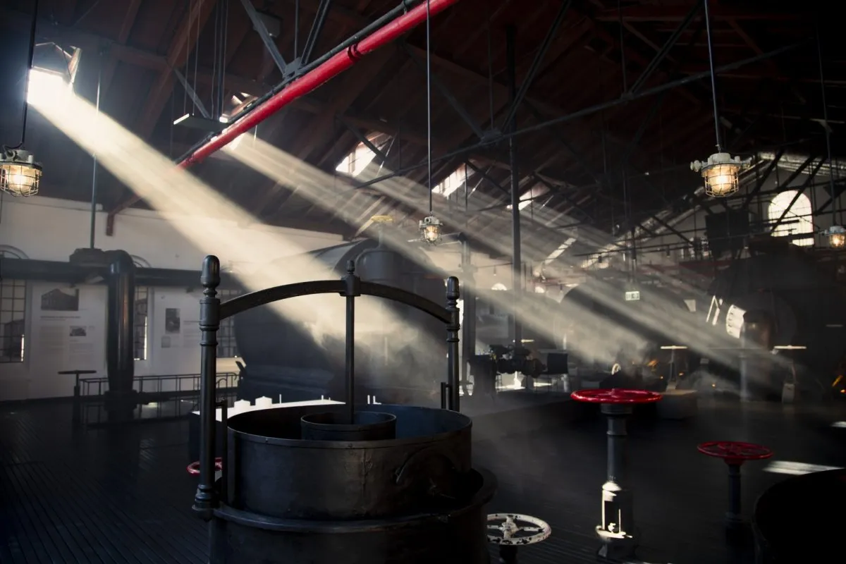 Sunlight beams through a large industrial hall with exposed red pipes and large machinery.