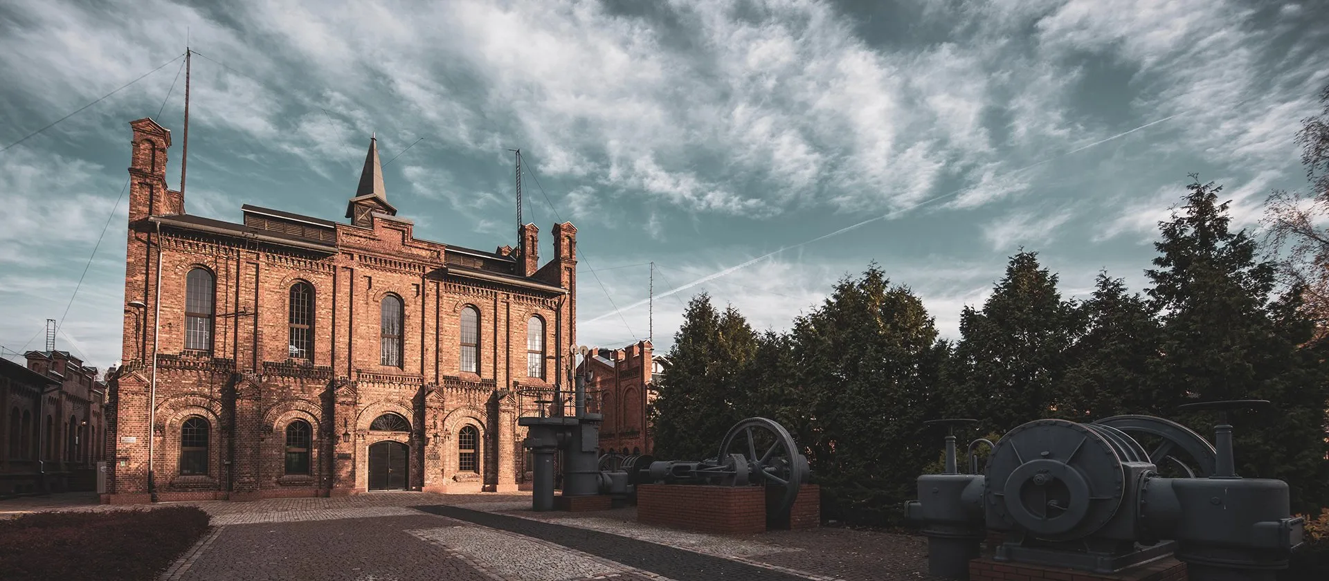 A historic brick building with a tower and arched windows, surrounded by industrial machinery and trees under a cloudy sky