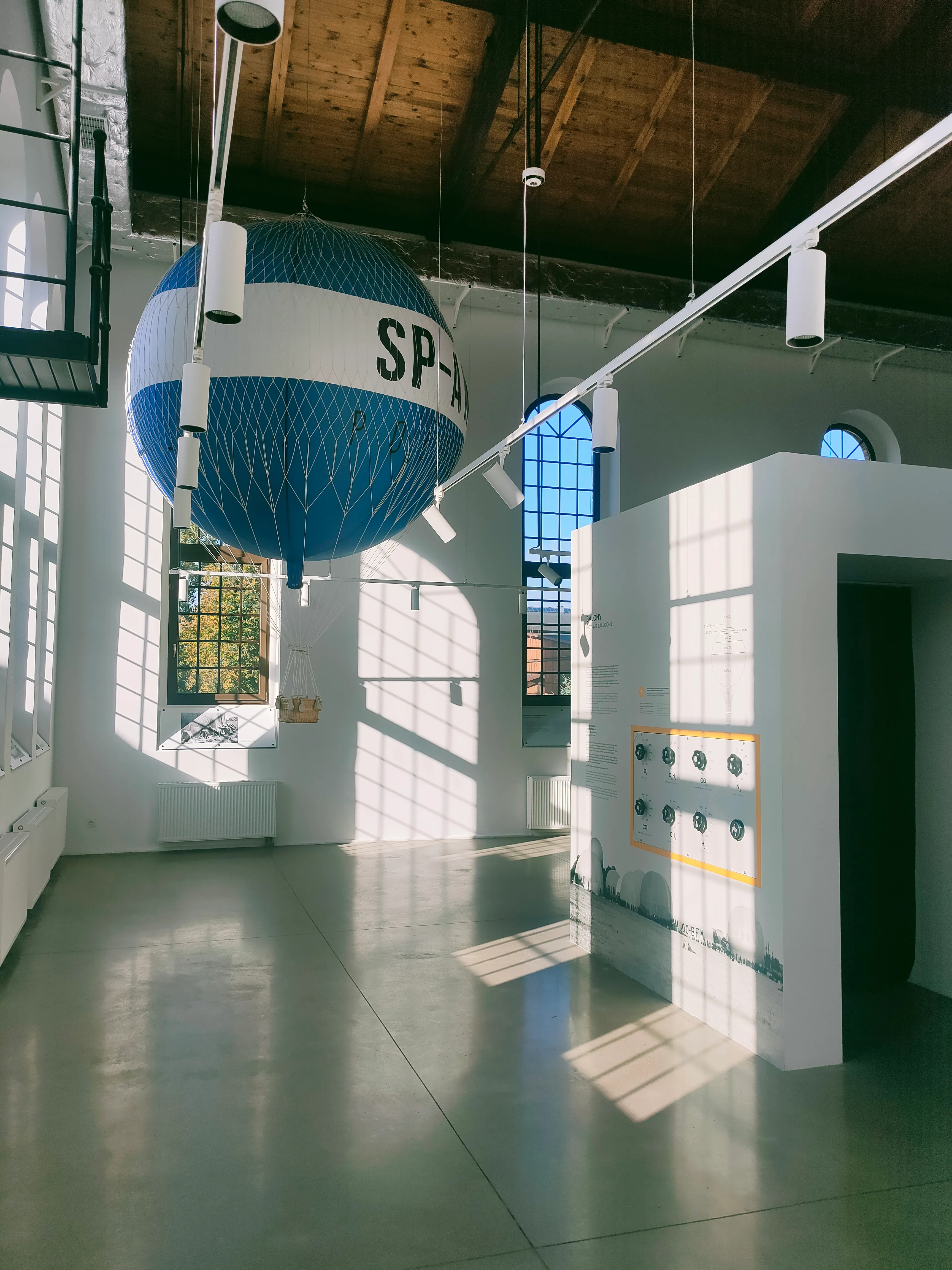 A blue and white spherical gas holder model hangs from the ceiling in a spacious industrial-style gallery with high ceilings and large windows.