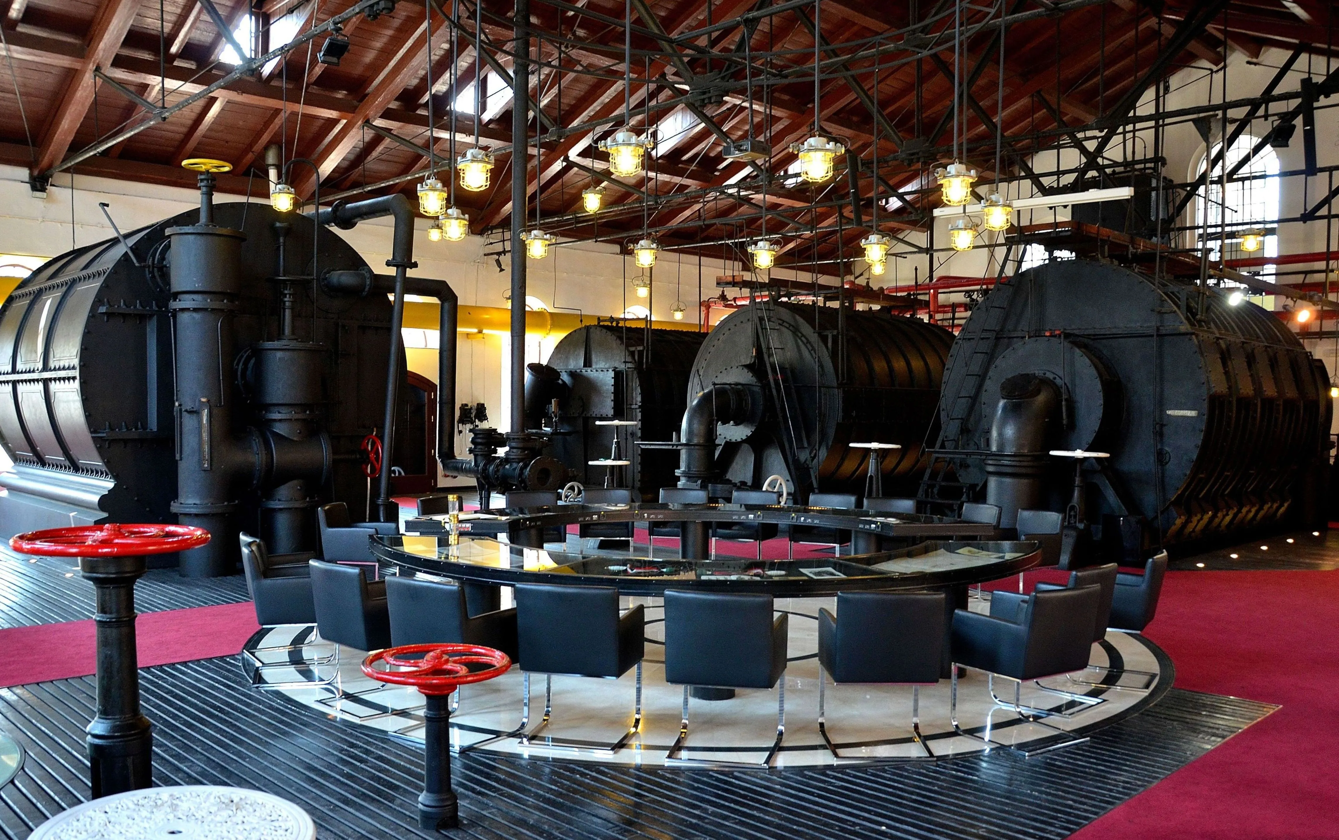 Interior of the Warsaw Gasworks Museum with large black gas holders, industrial machinery, and a circular seating area with red flooring and black chairs.