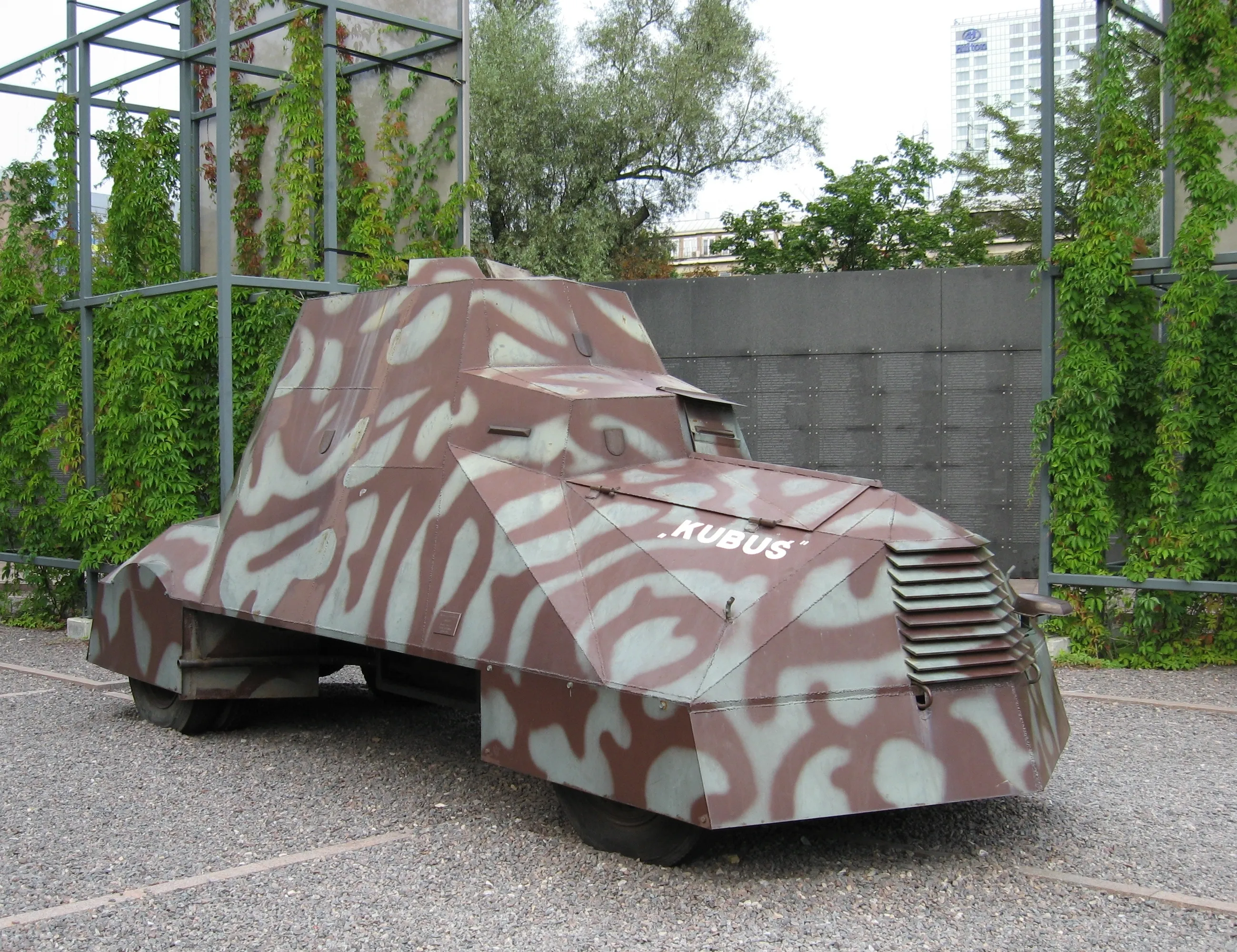 Vehículo militar camuflado con el texto 'KUBUS' en la parte delantera, al aire libre cerca de vegetación y un edificio