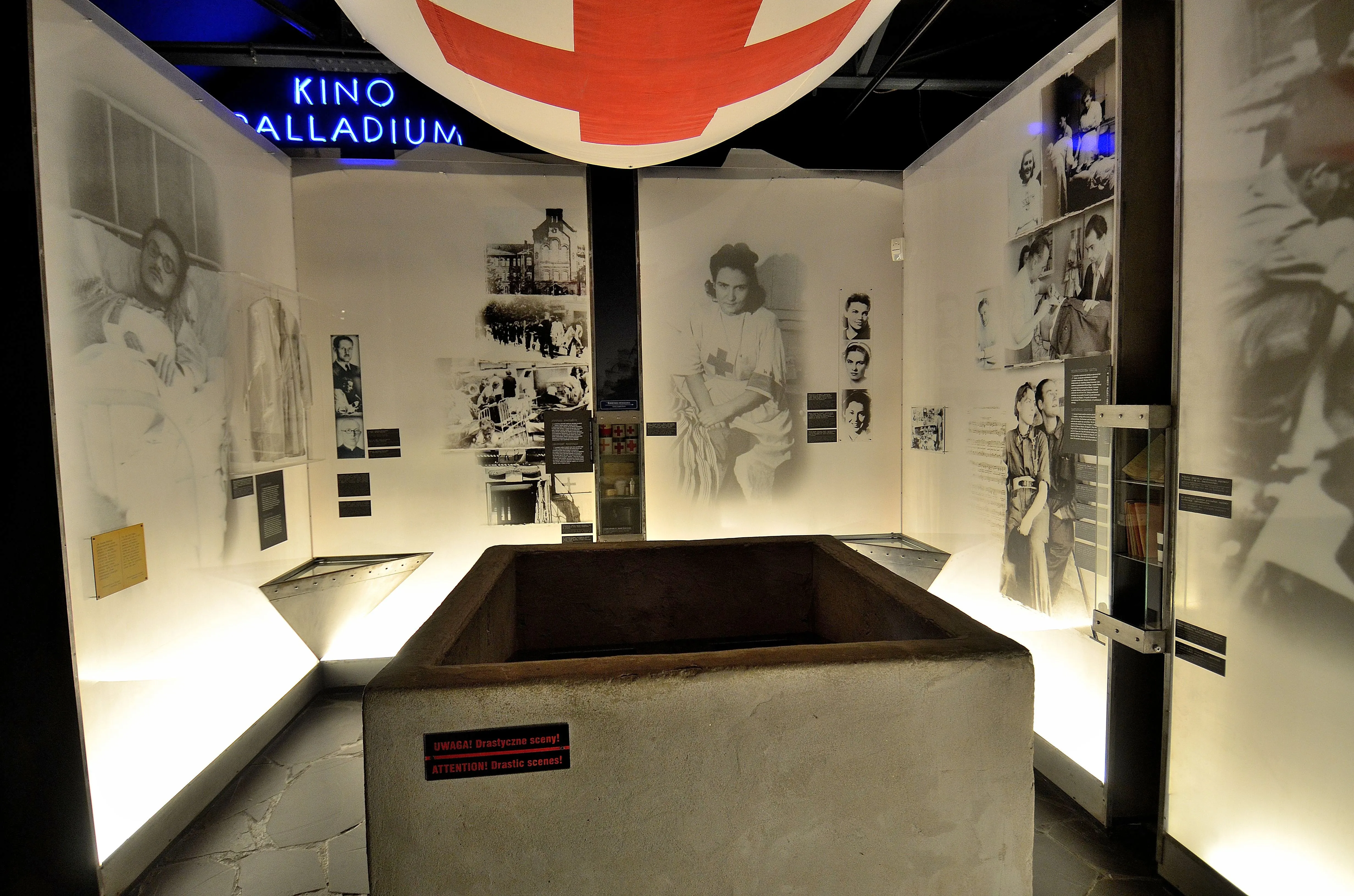 Exhibición interior del Museo del Alzamiento de Varsovia con una cama de hospital, fotos en blanco y negro en las paredes y un emblema rojo y blanco colgado del techo