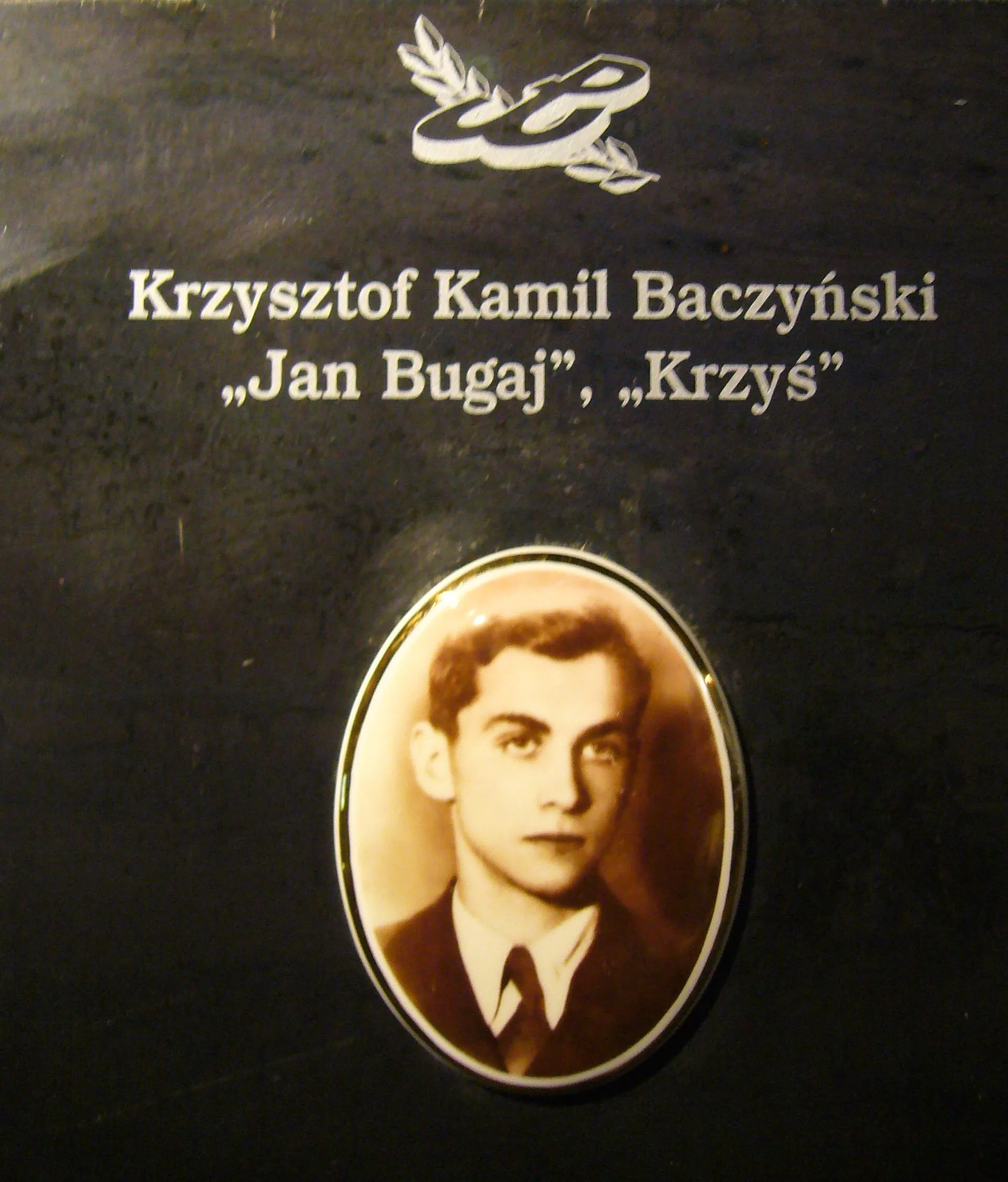 Een zwarte plaquette met een cirkelvormig portret van een jonge man en tekst die hem identificeert als Krzysztof Kamil Baczyński, met de pseudoniemen Jan Bugaj en Krzyś.