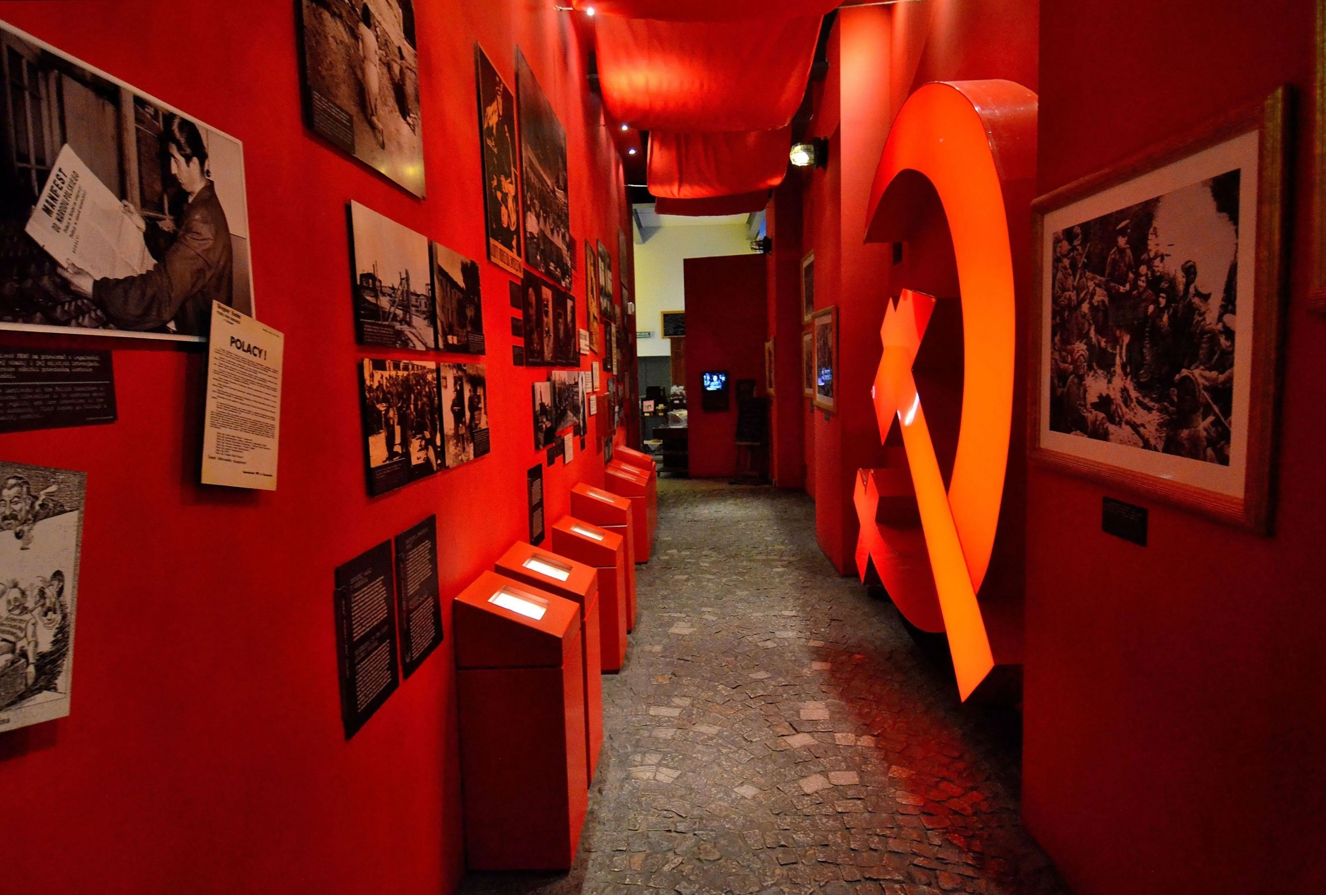 Pasillo de museo de paredes rojas con fotografías históricas, vitrinas y un gran elemento escultórico rojo