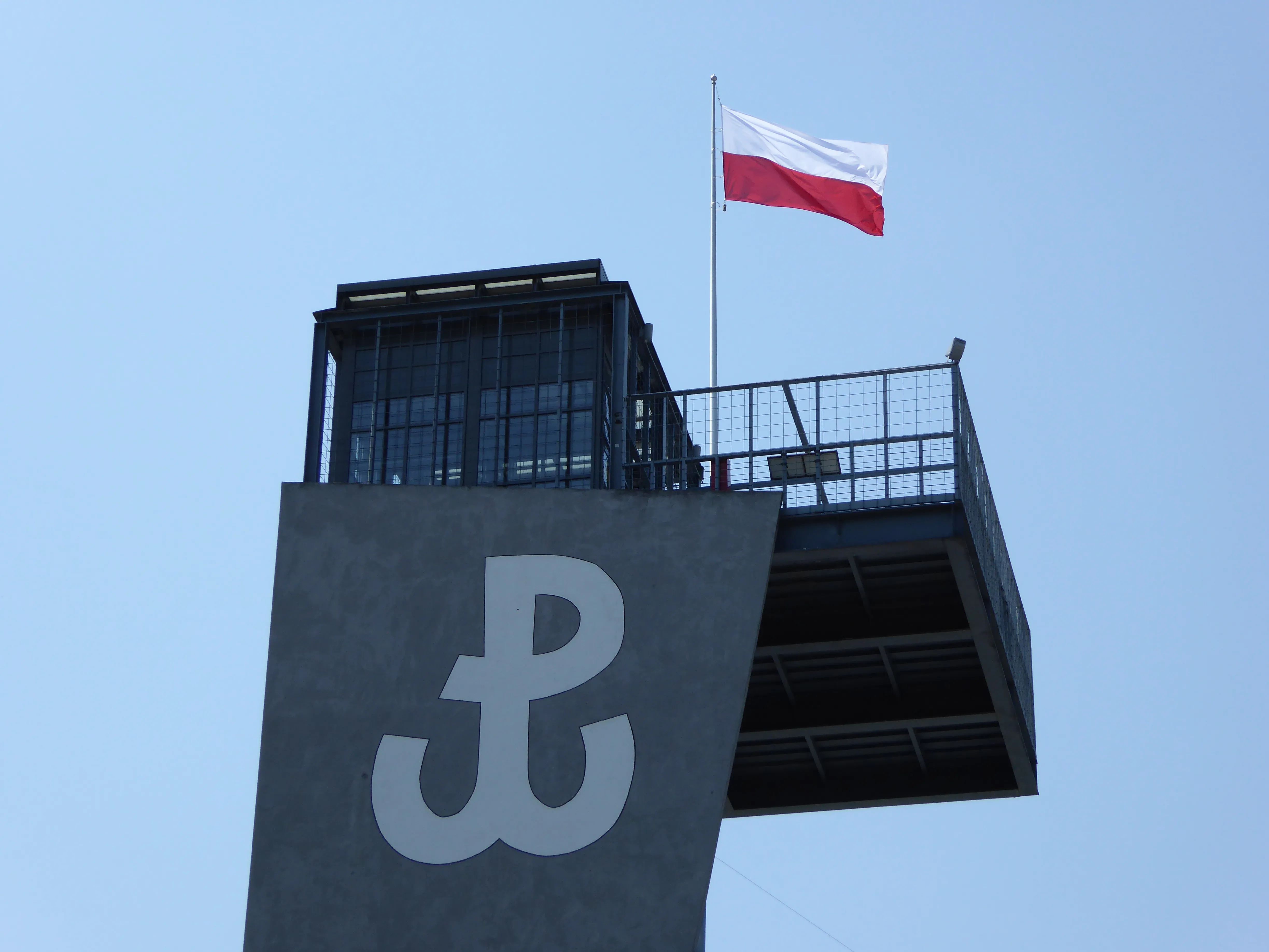 Torre de observación con un gran símbolo blanco 'P' y una bandera polaca en la parte superior contra un cielo azul
