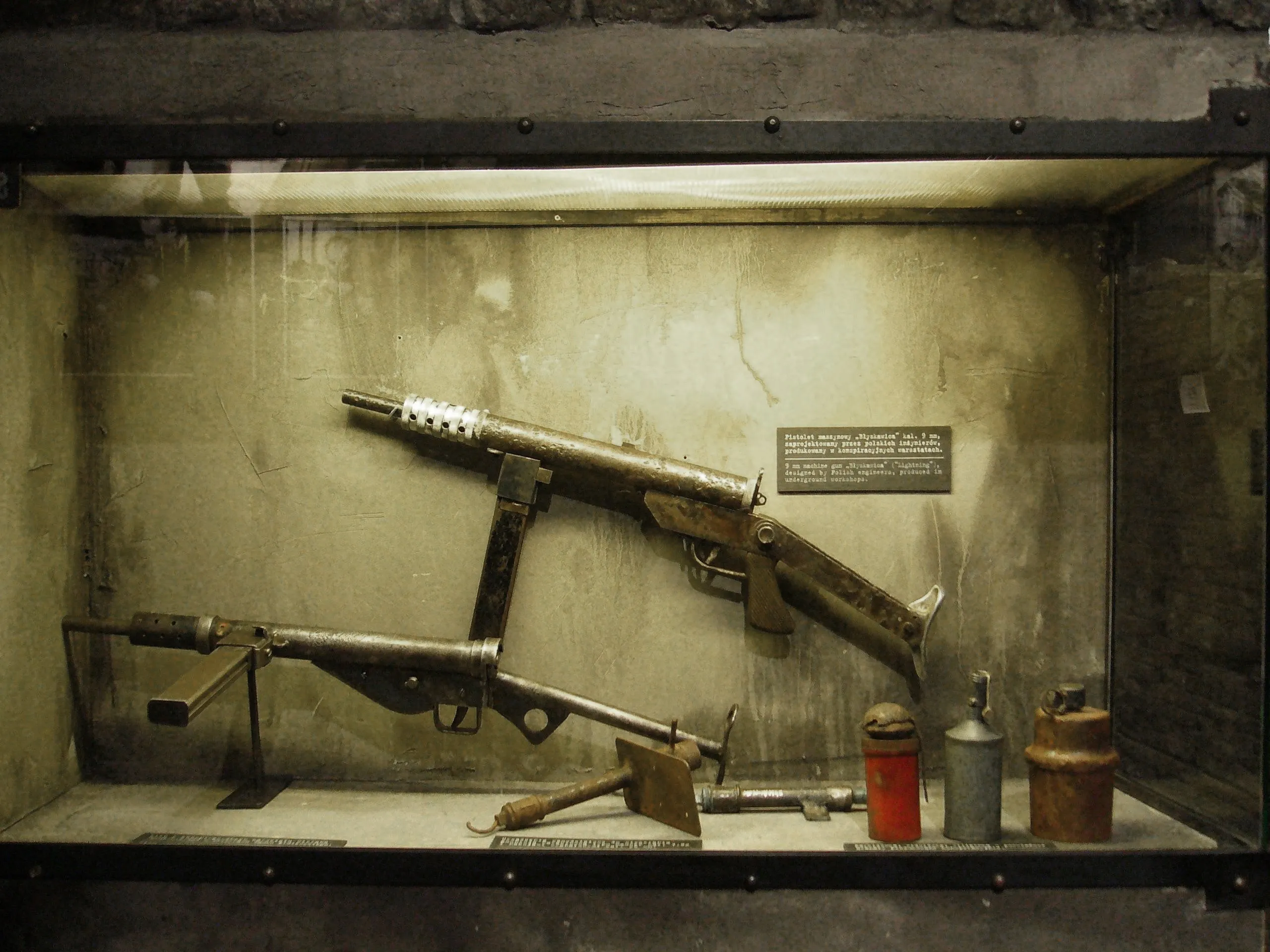 vitrina con armas insurgentes que incluyen una ametralladora Błyskawica y otras armas de fuego, un bidón rojo y un contenedor metálico
