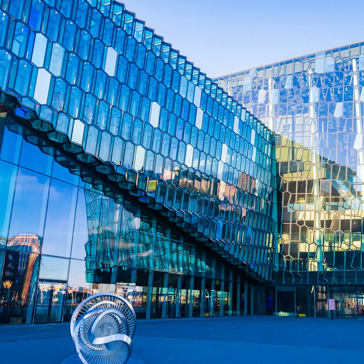 Edificio moderno con fachada de cristal hexagonal azul y una escultura metálica en espiral en primer plano al atardecer
