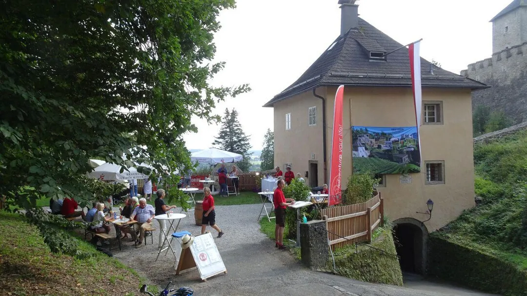 Vista exterior del edificio Georgsturm con techo cónico, entrada de túnel, banderas rojas y visitantes sentados en mesas al aire libre en una zona ajardinada.