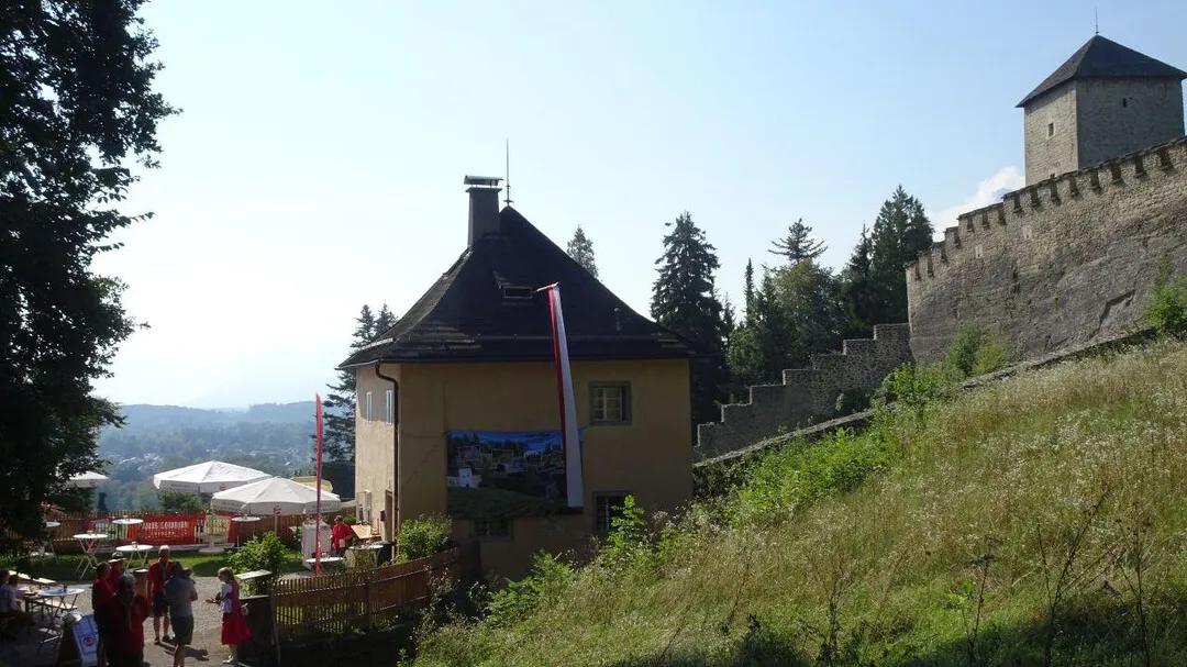 Vista exterior de un edificio histórico con un tejado cónico oscuro junto a un muro de fortificación de piedra y una torre cuadrada en una ladera cubierta de hierba, con visitantes caminando cerca