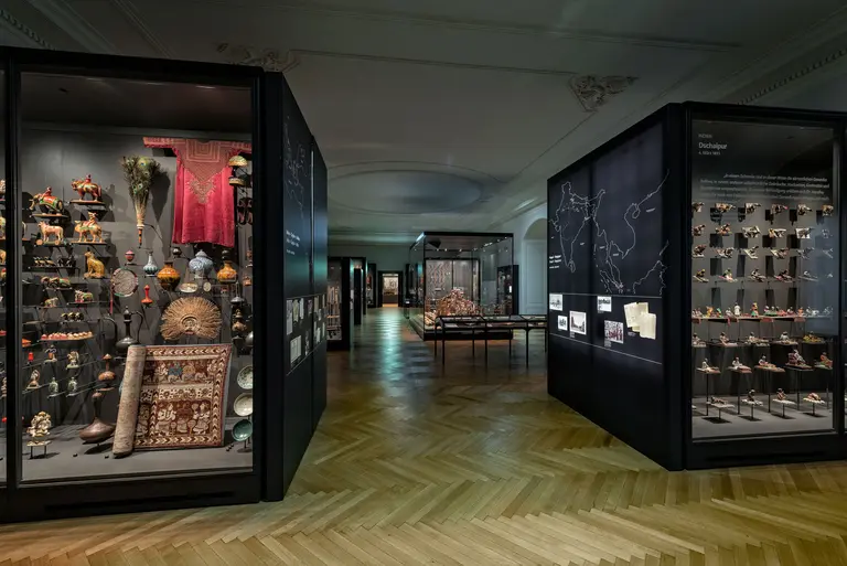 A museum gallery with glass display cases filled with cultural artifacts and a wooden herringbone floor