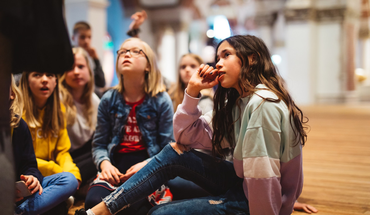 Group of children sitting on wooden floor in museum hall, looking up at exhibition. Warm lighting, high ceilings visible.