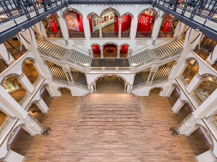 Grand atrium of Wereldmuseum Amsterdam with multi-level staircases, white arches, and red banners displaying words including LOVE