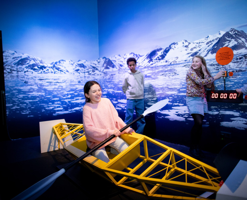 Three people interact with a yellow boat structure in front of a mural depicting snowy mountains and water, with a digital timer display