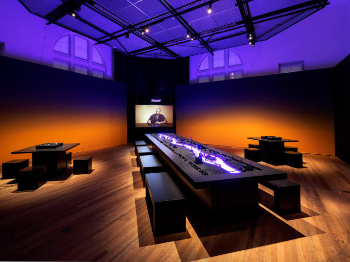 Modern exhibition hall with long display table illuminated in blue, wooden floors, dark walls with orange lighting, and viewing benches at Wereldmuseum Amsterdam.