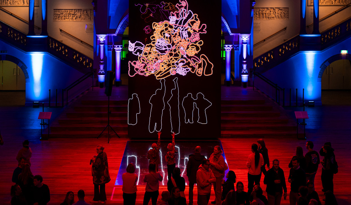 Dramatically lit museum interior with blue and red lighting featuring a large illuminated art installation with abstract figures and gathered visitors