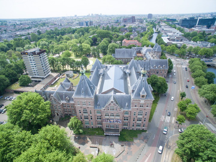 Aerial view of Wereldmuseum Amsterdam's historic building with gray-tiled towers, red-brick facade, surrounded by lush green trees and near a waterway.