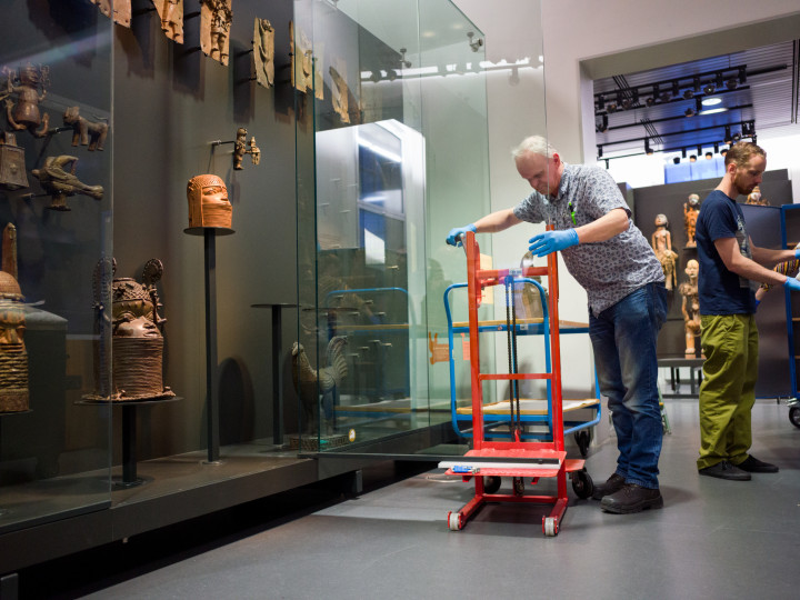 Staff moving a red dolly near glass display cases containing cultural artifacts and sculptures in a museum exhibition space.