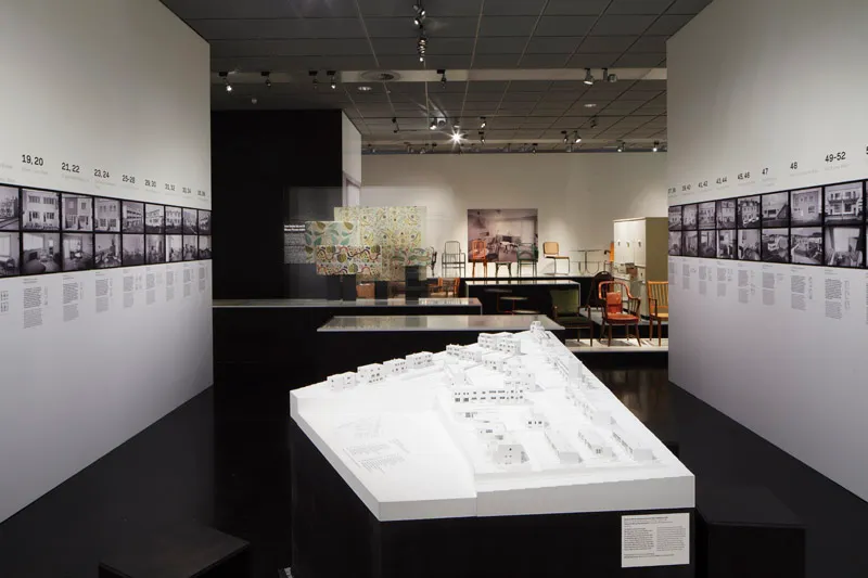 Ekspozycja z modelami architektonicznymi, fotografiami i meblami w galerii z białymi ścianami i oświetleniem punktowym