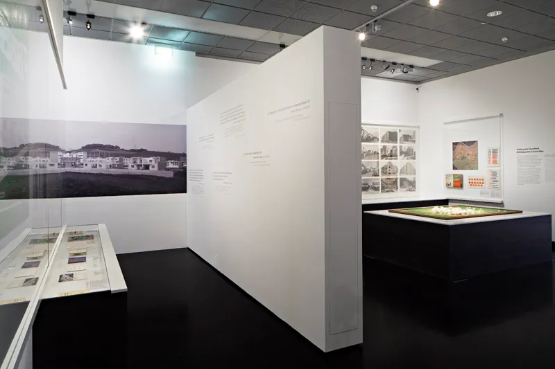 Sala wystawowa z białymi ścianami, dużym historycznym zdjęciem osiedla mieszkaniowego, gablotami i centralnym modelem architektonicznym pod reflektorami.