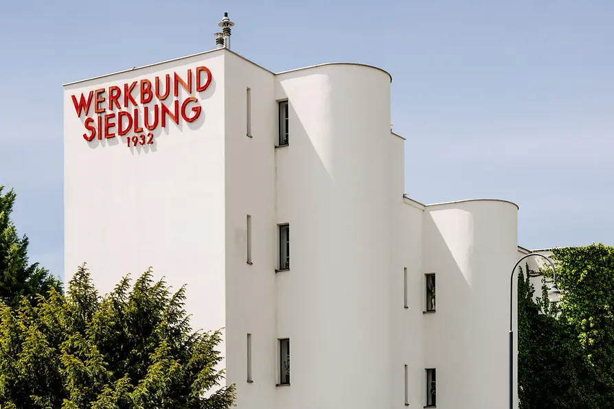 Biały modernistyczny budynek z czerwonym napisem 'WERKBUND SIEDLUNG 1932' na fasadzie, otoczony zielonymi drzewami pod błękitnym niebem
