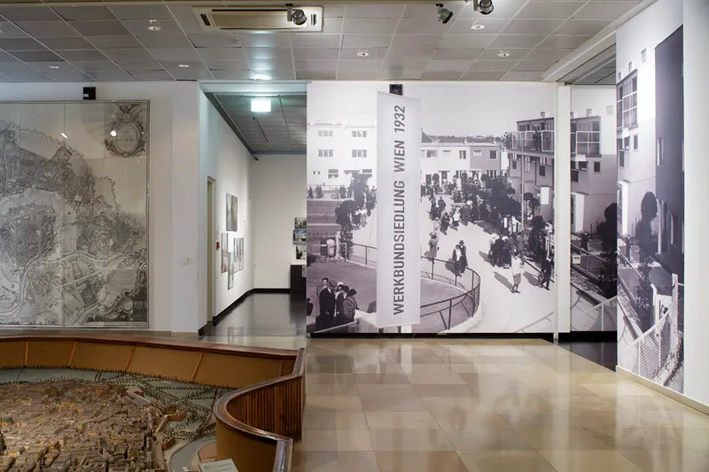 Una galería de museo con grandes fotografías históricas de la Urbanización Werkbund, que incluye una imagen destacada en blanco y negro del complejo de viviendas de 1932 y gente en la calle.