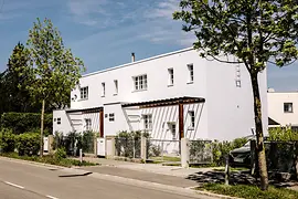 biały modernistyczny budynek z płaskim dachem i dużymi oknami, otoczony drzewami i ulicą