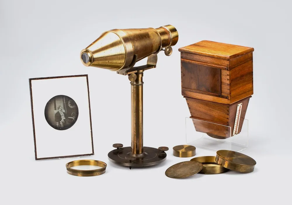 Historic photographic equipment including a brass camera obscura, a wooden box camera, a framed photograph, and lens components