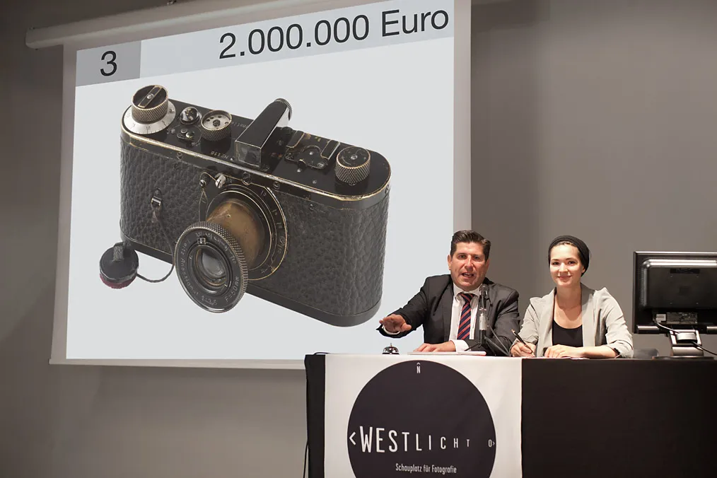 A vintage camera displayed on a screen with a price tag of 2,000,000 Euro, with two people seated at a table in front of the screen.