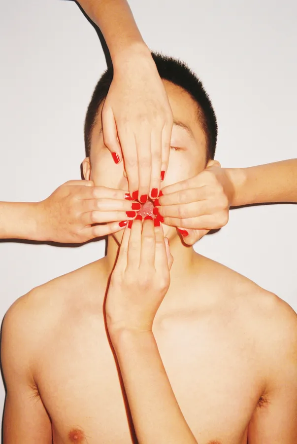 A person with a bare chest and closed eyes has their face covered by several hands with red nail polish against a plain white background.