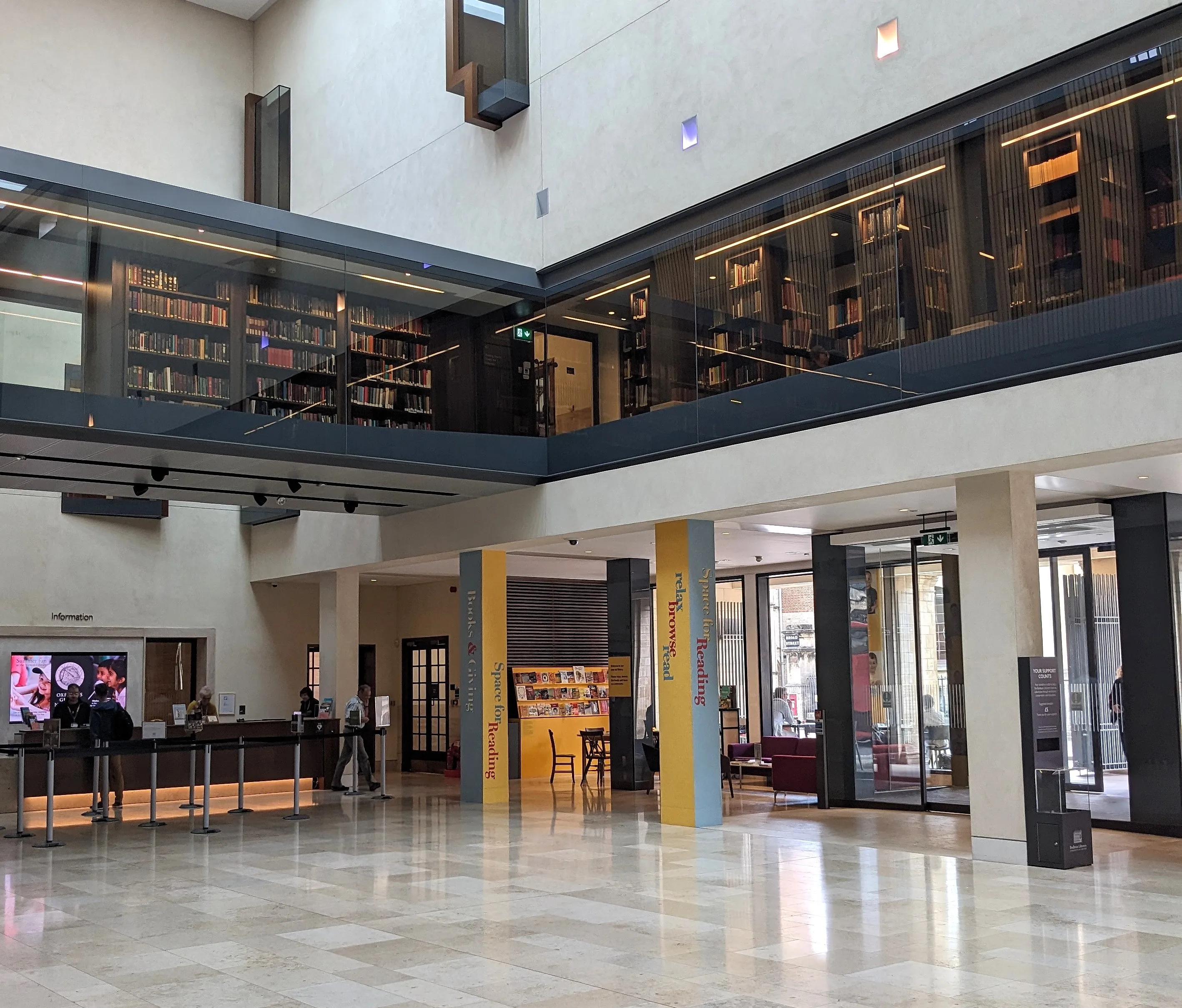 Spacious interior hall of the Weston Library with a mezzanine level lined with bookshelves behind glass, a ground-floor reception desk, and large entrance doors