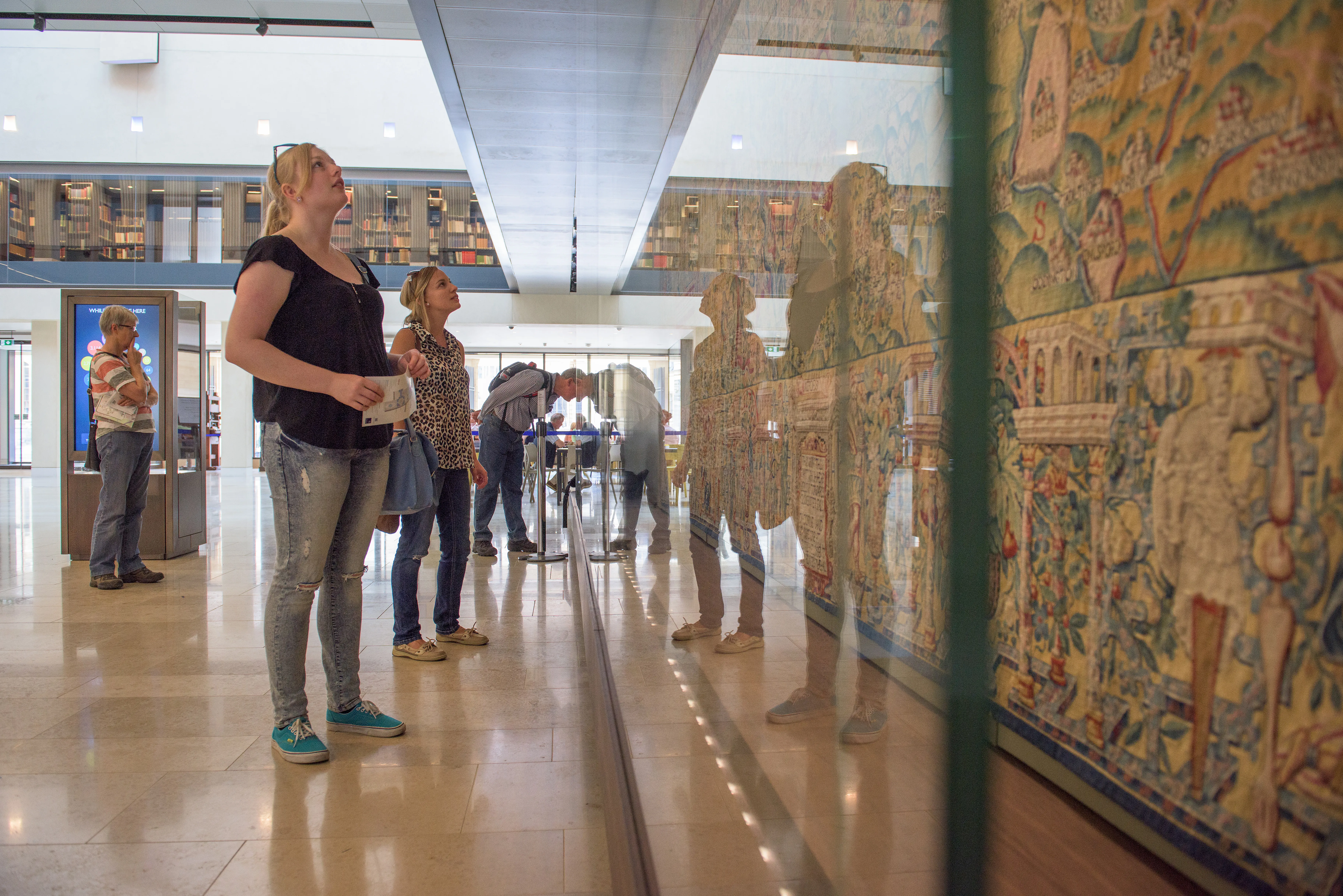 Visitors standing in Blackwell Hall at the Weston Library viewing a large colorful tapestry displayed behind a glass case
