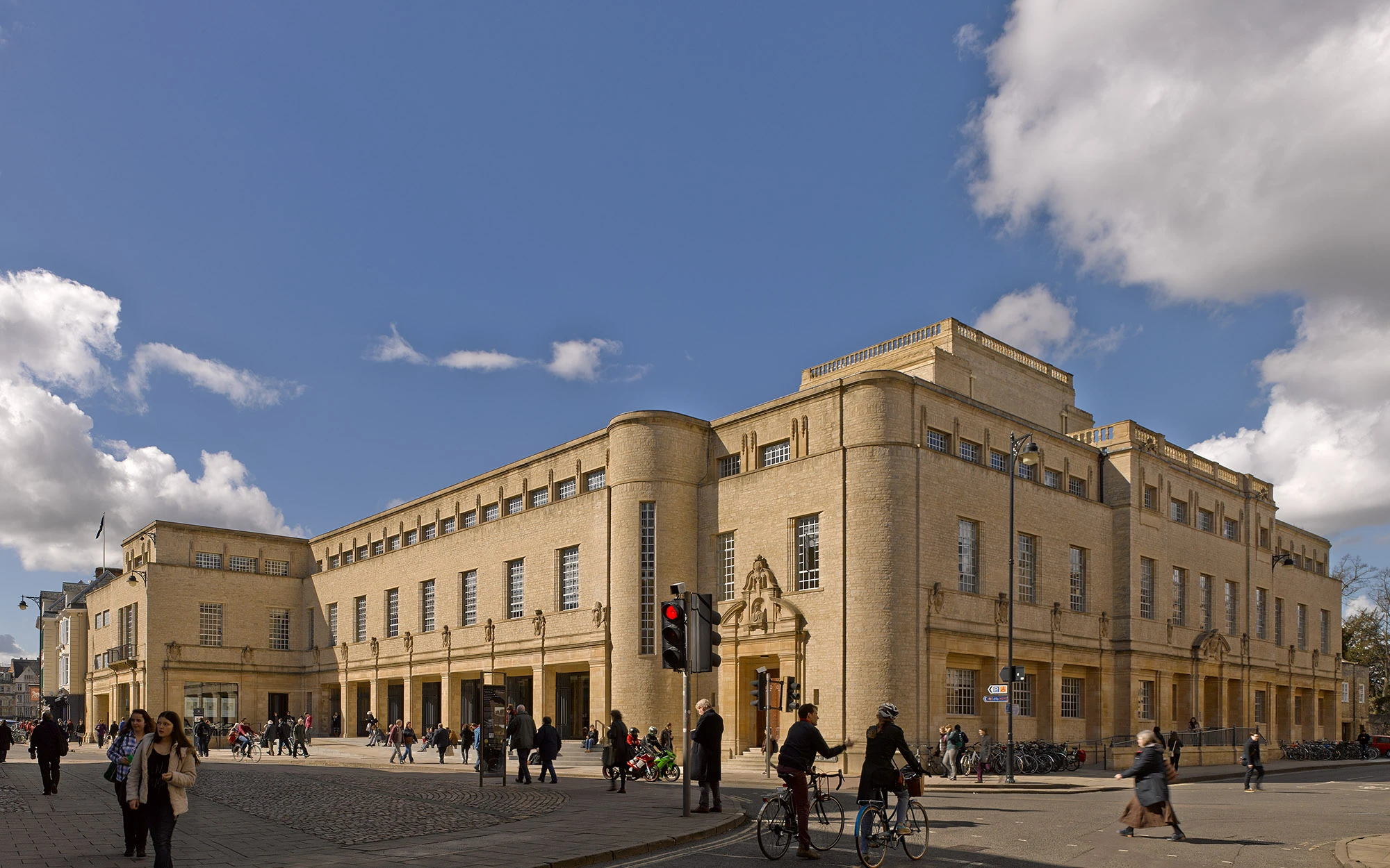 Wide exterior view of the Weston Library, a large light-colored stone building with Art Deco architecture, with people walking and cycling in front under a blue sky with scattered clouds