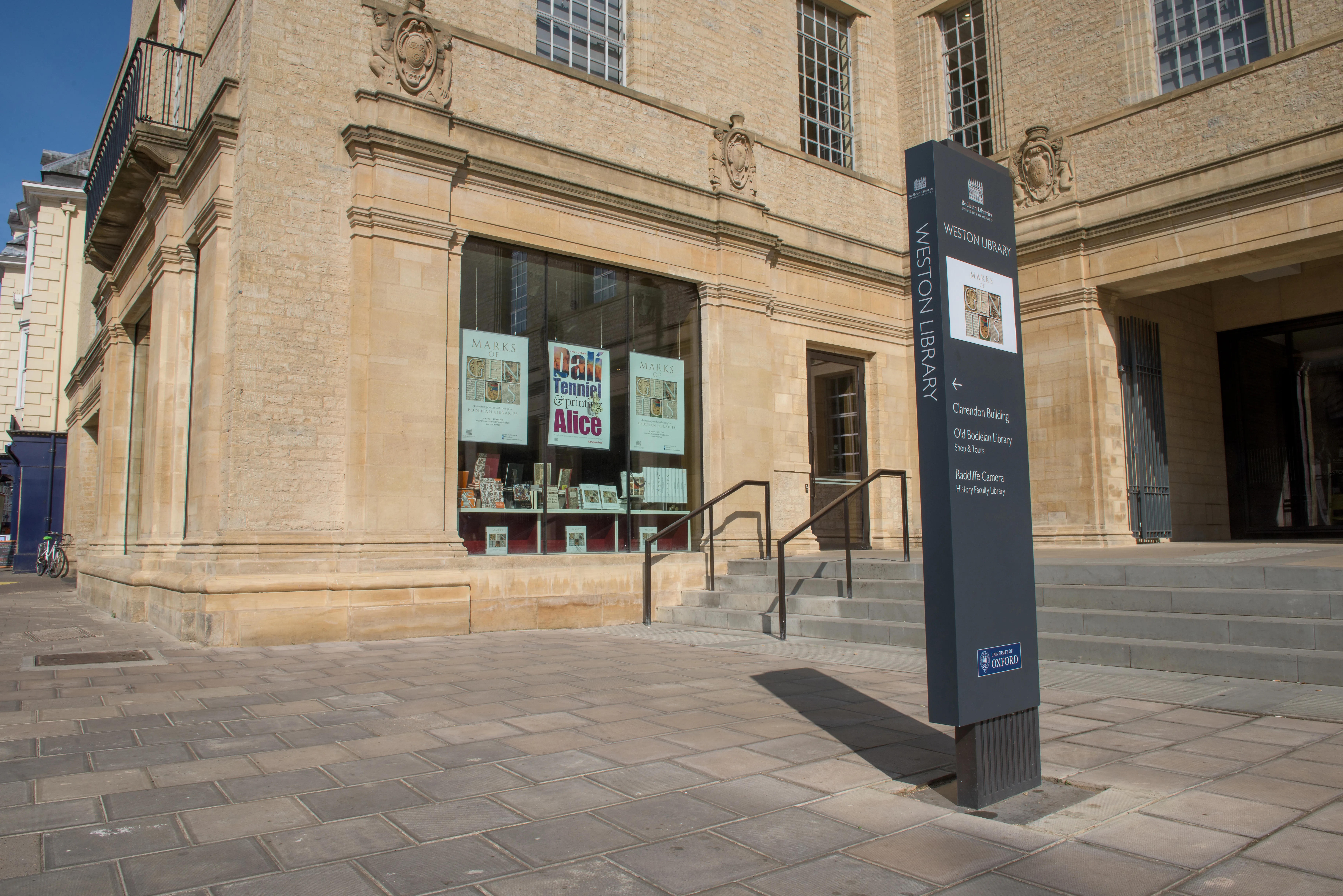 Stone facade of the Weston Library with large display windows showing exhibition posters and a tall dark signpost reading Weston Library in the foreground