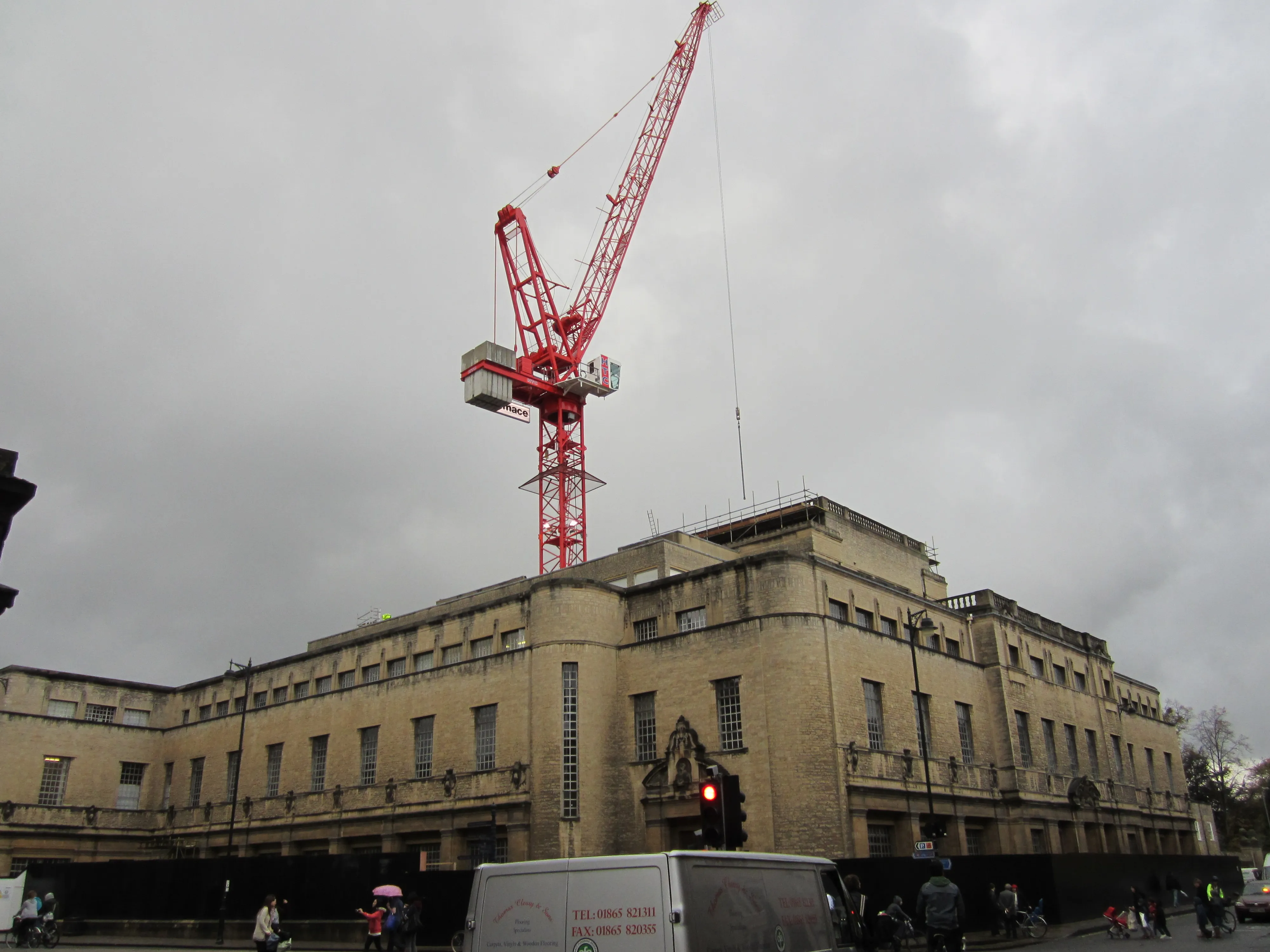 Beige stone Art Deco building with a tall red construction crane towering above it under an overcast gray sky, with a white van and pedestrians at street level