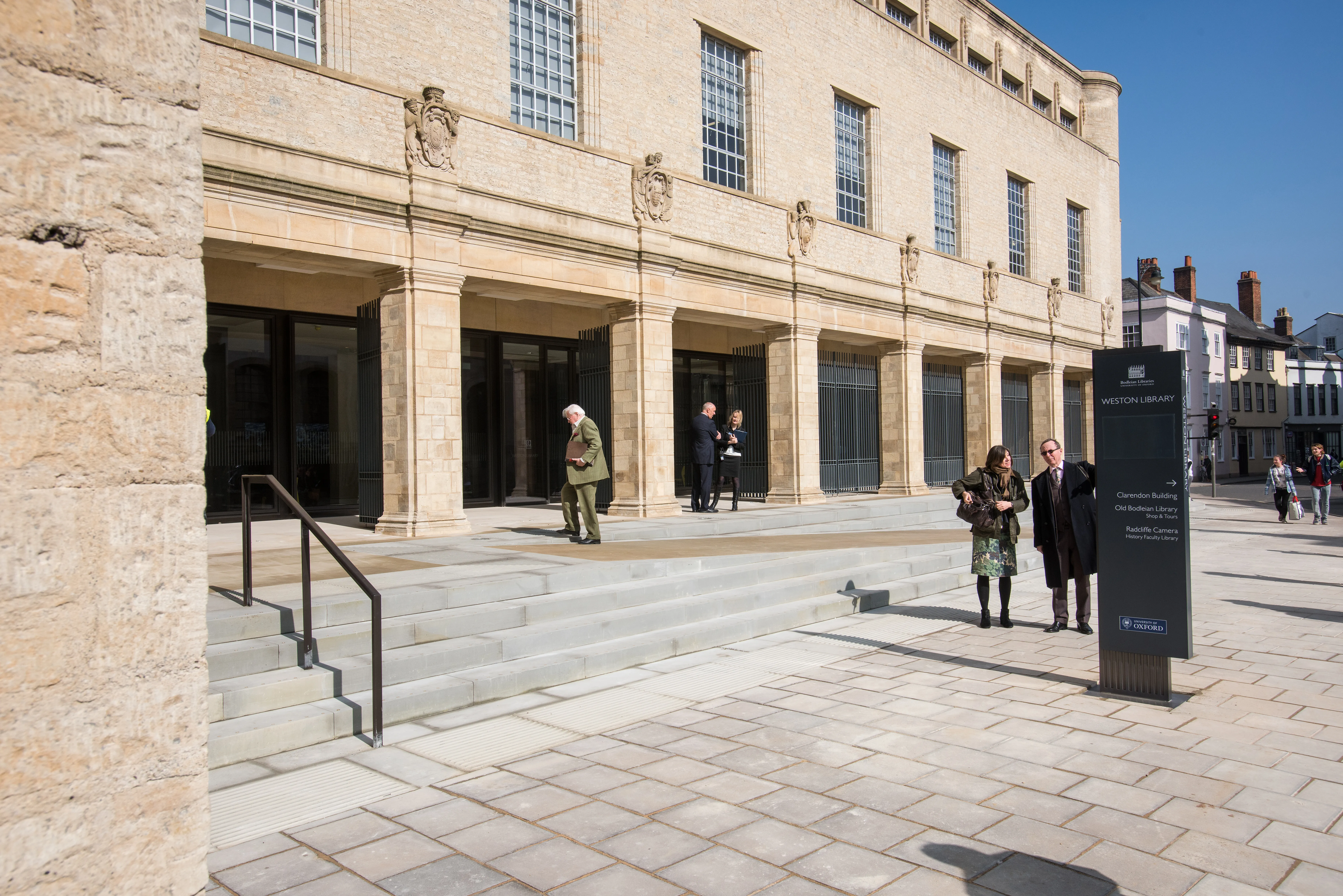 Stone facade of the Weston Library with tall windows, columned entrance with steps, and visitors standing near a black information sign