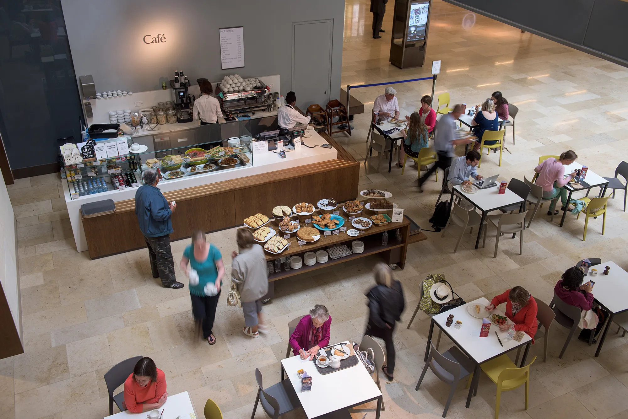 Overhead view of the Blackwell Hall café inside Weston Library, showing a food counter, display tables with pastries, and visitors seated at white tables