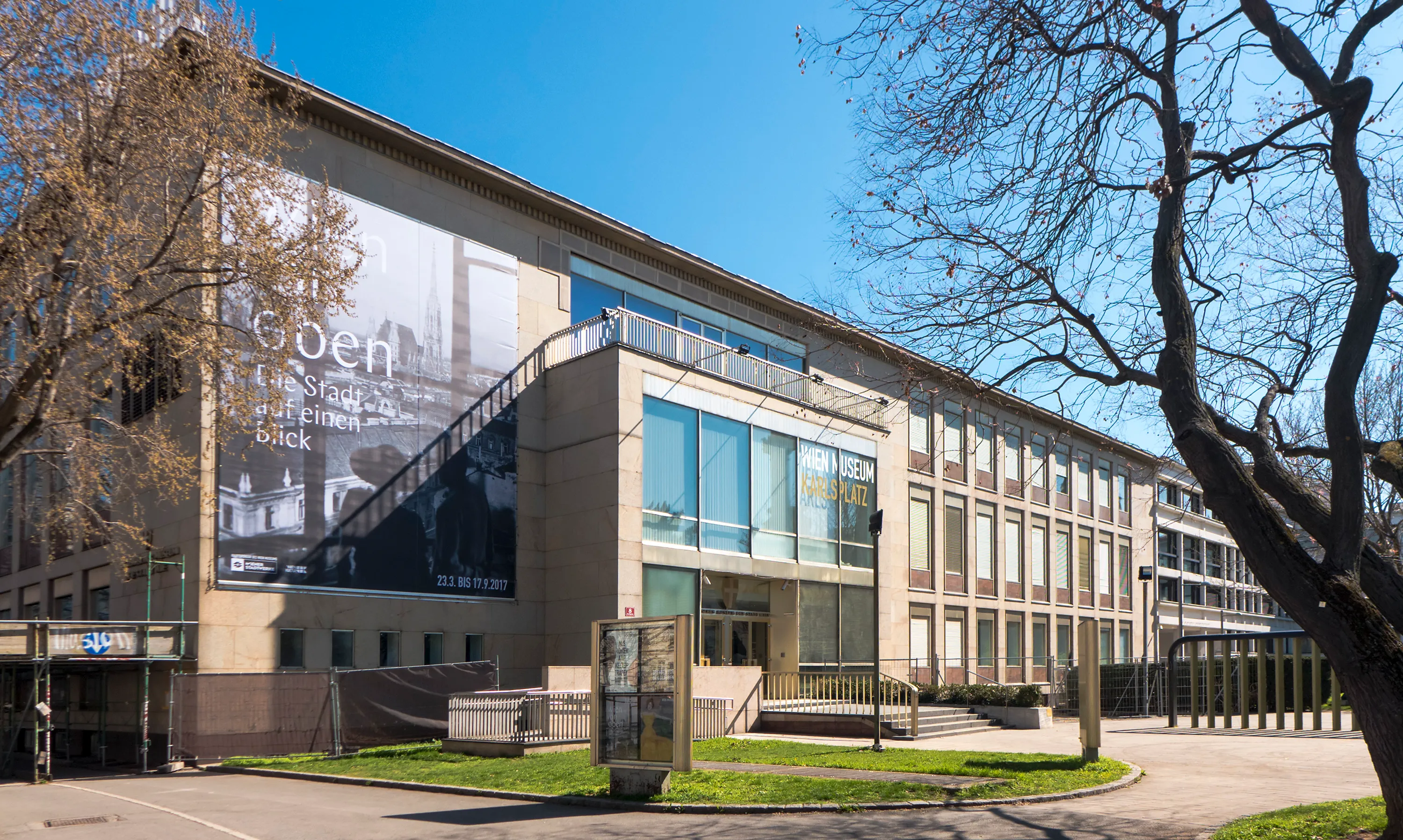 Wien Museum-gebouw met een grote zwart-witte historische foto op de gevel, een moderne glazen ingang en kale bomen ervoor onder een helderblauwe hemel