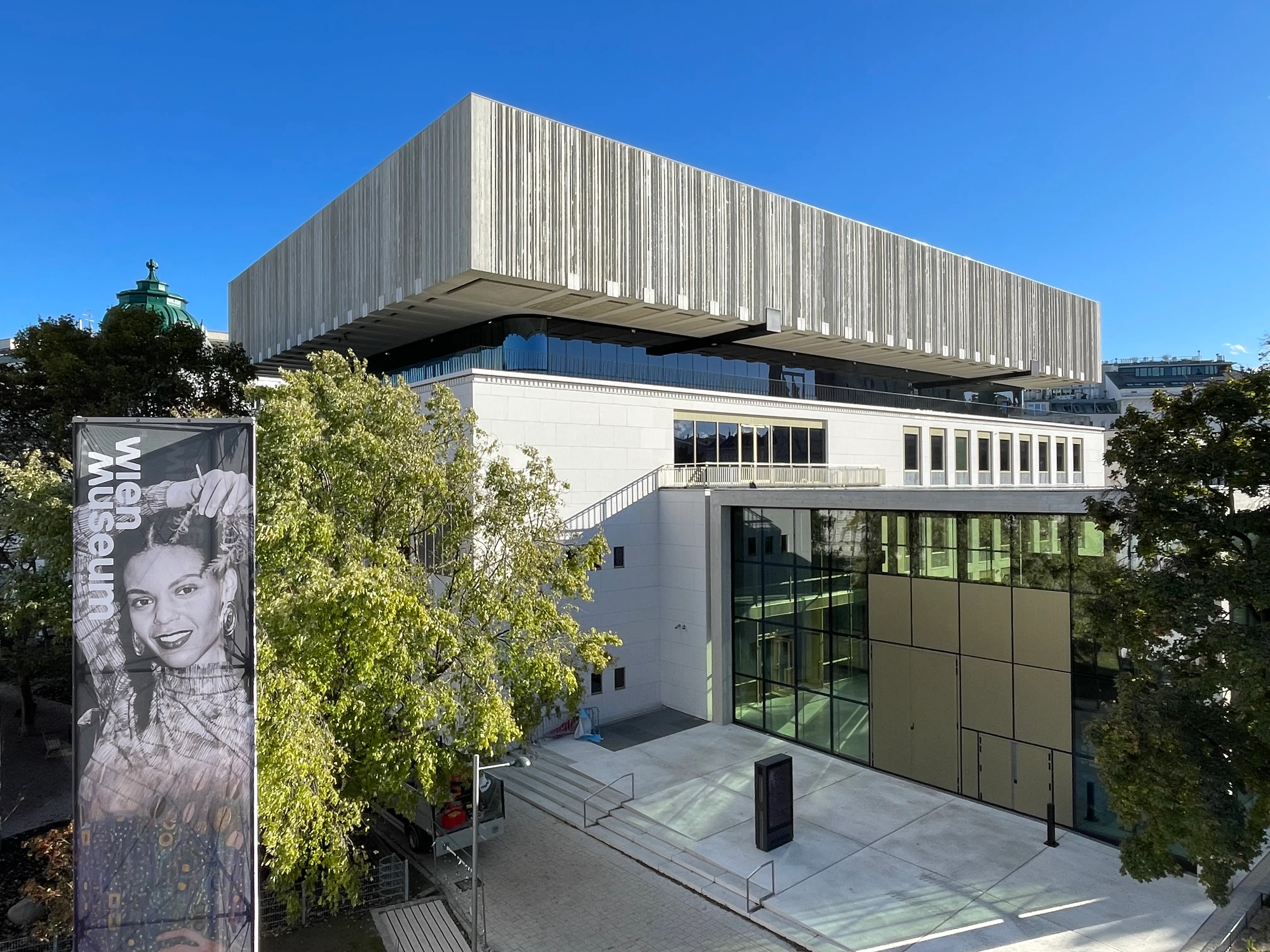 Modern gebouw met een betonnen gevel en glazen ingang, omringd door bomen onder een blauwe lucht