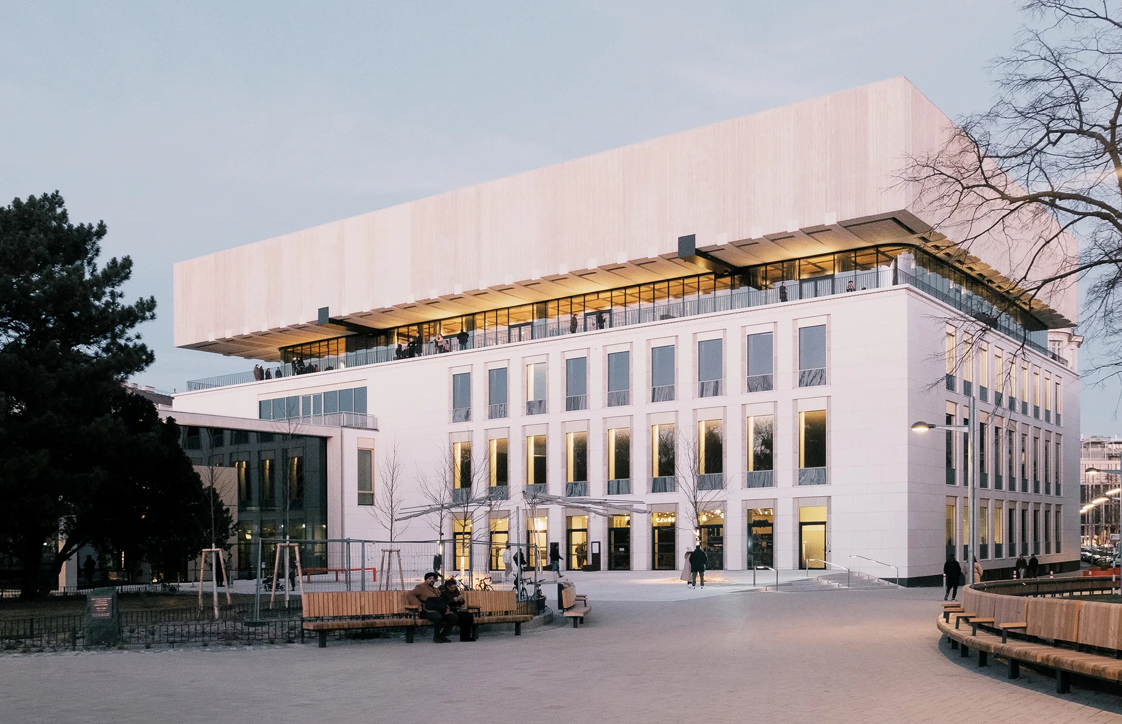 Modern white building with a large overhanging roof and illuminated windows, set against a dusky sky with trees and benches in the foreground.